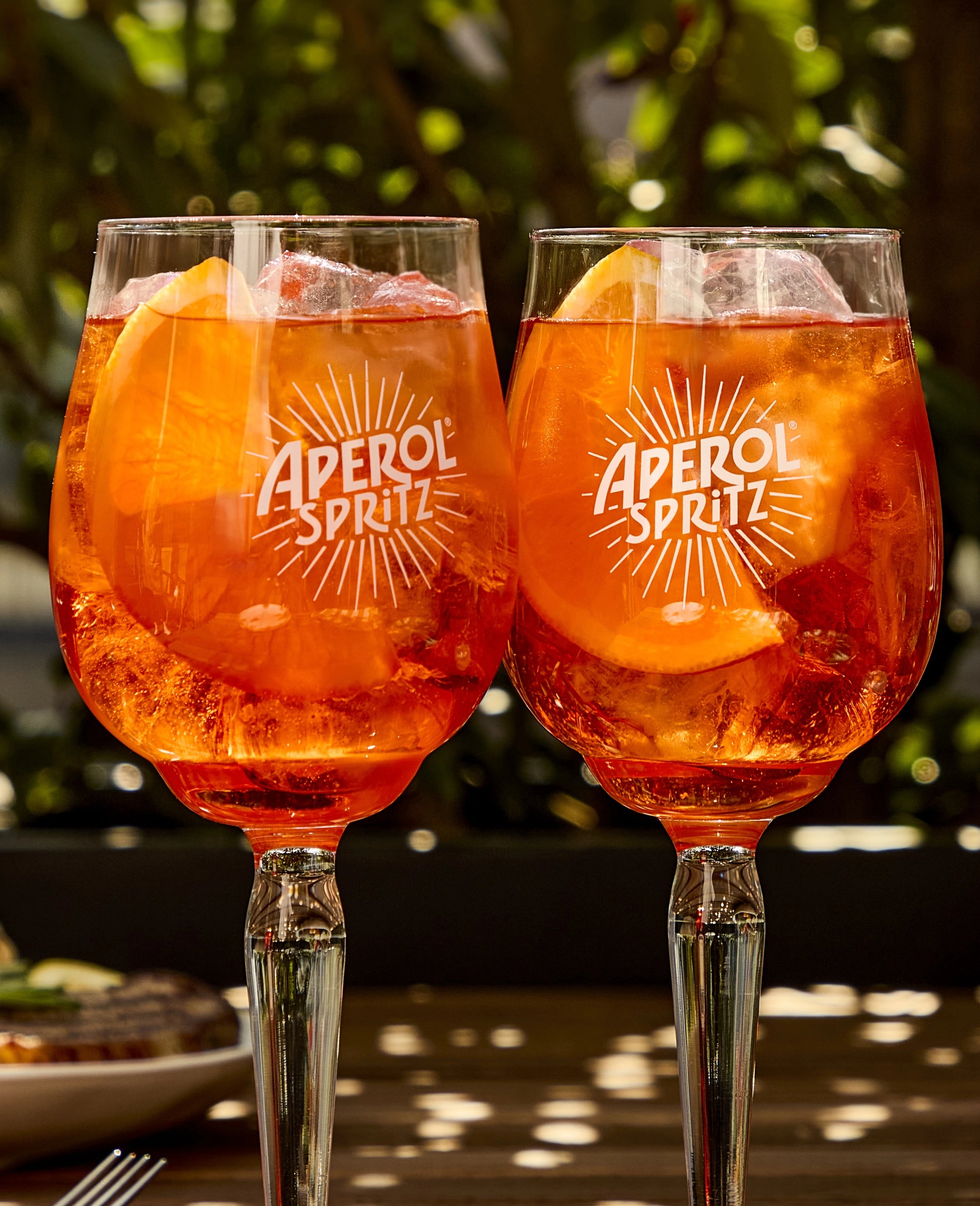 Two Aperol Spritz glasses filled with ice and orange slices, served outdoors on a sunlit wooden table