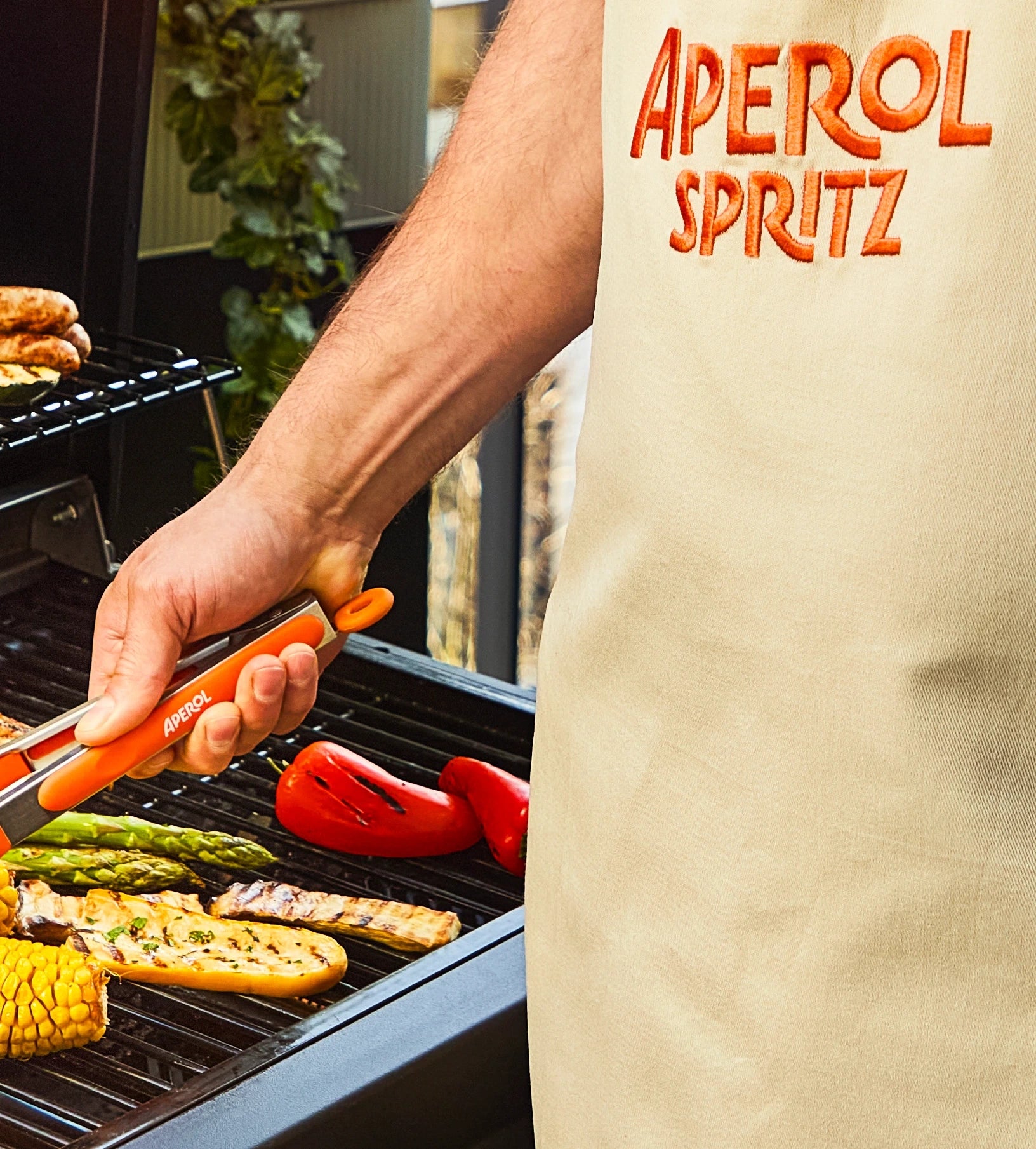 Person grilling vegetables wearing an Aperol Spritz apron and using branded tongs during outdoor barbecue