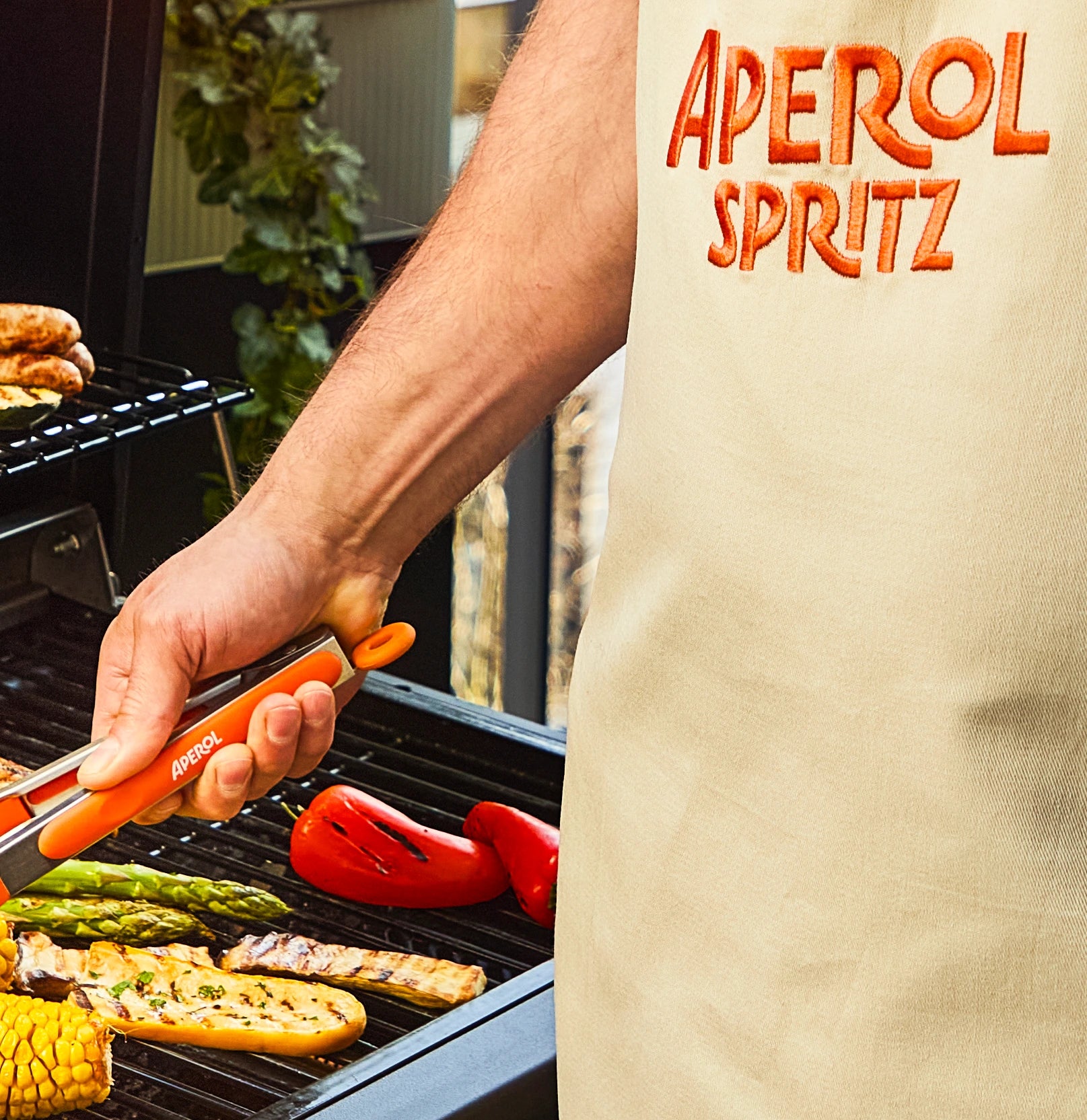 Person grilling vegetables wearing an Aperol Spritz apron and using branded tongs during outdoor barbecue