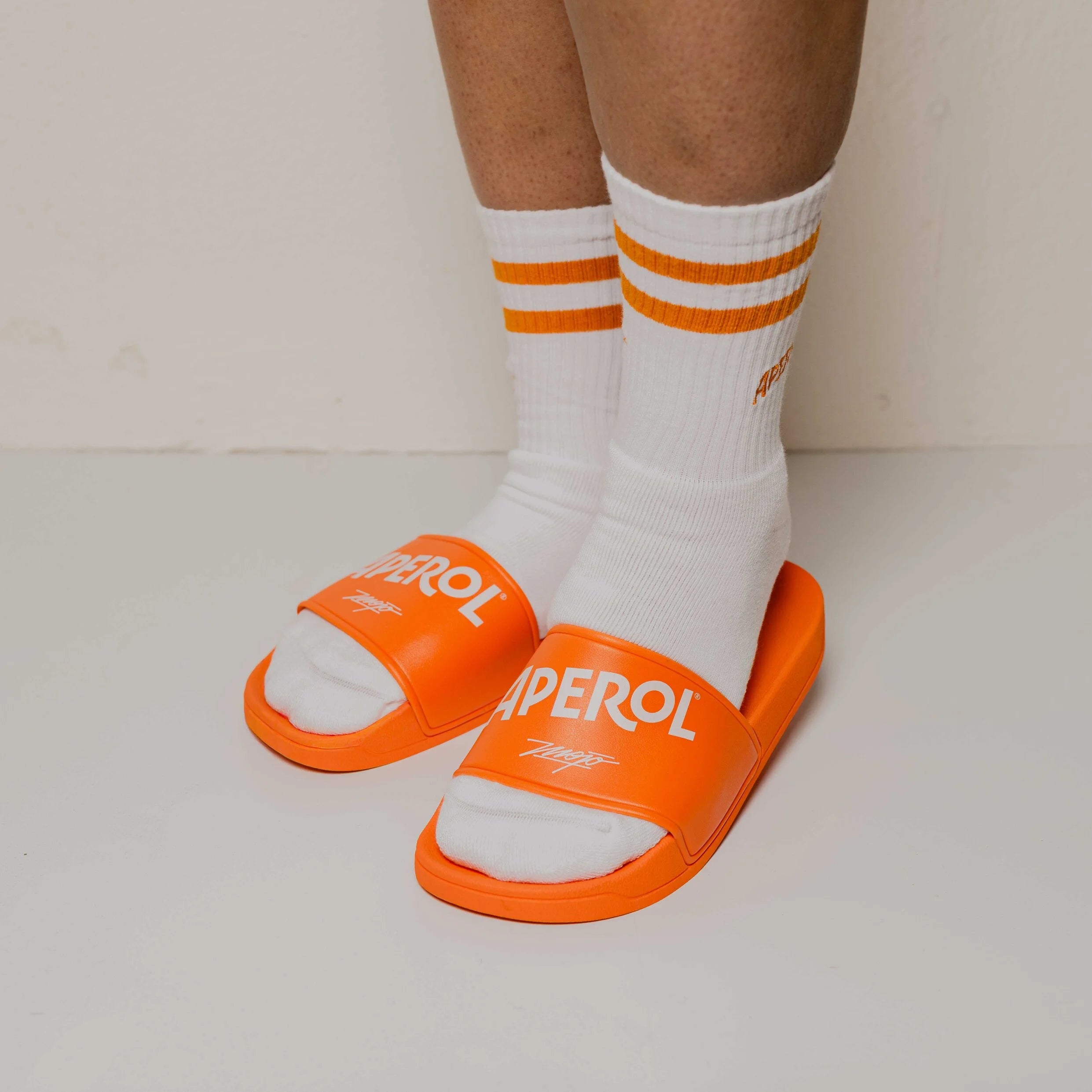 Orange slides with 'Aperol' branding worn with white socks featuring orange stripes on a light background