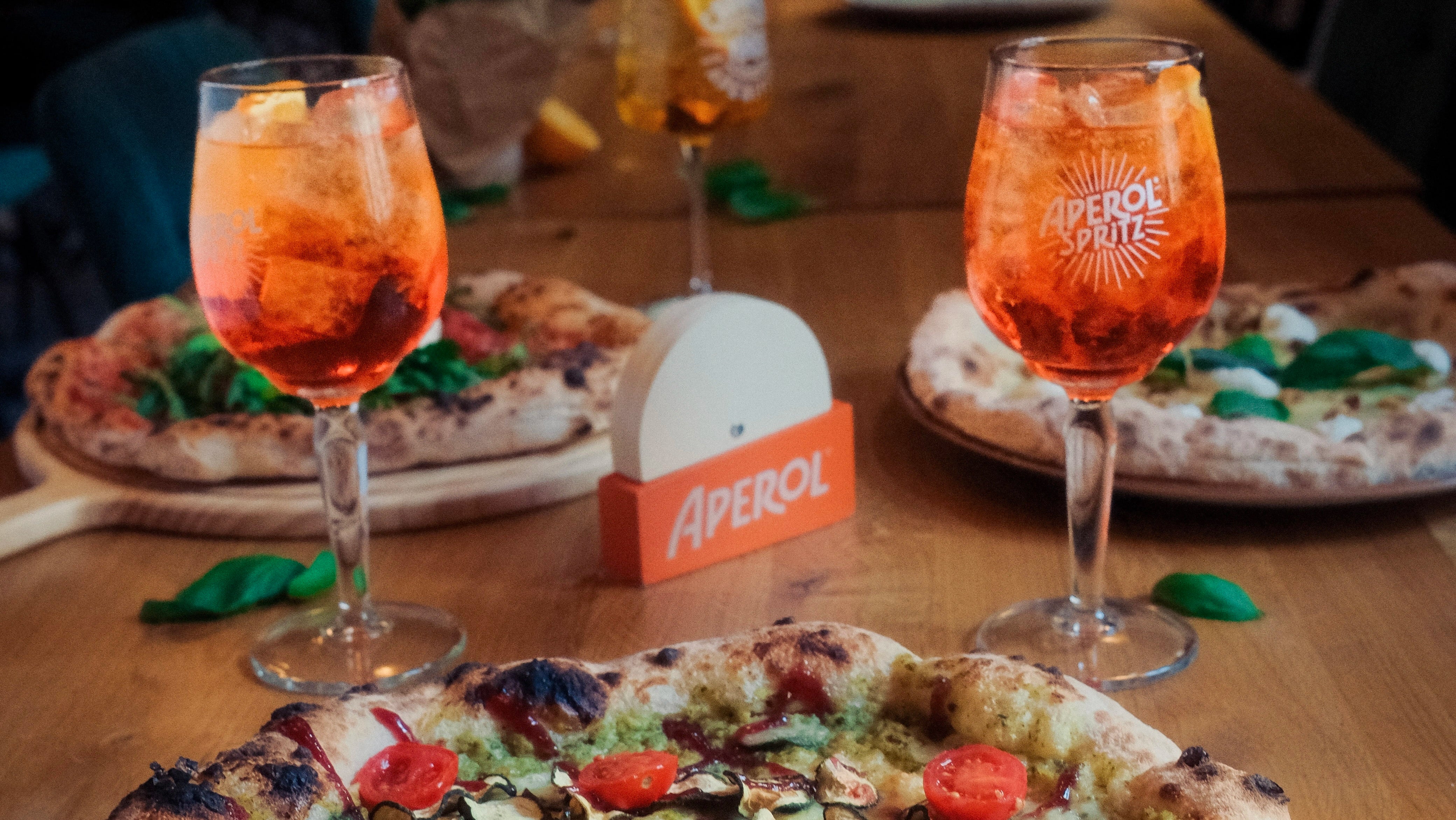 Two glasses of Aperol Spritz with pizzas on a wooden table, featuring Aperol branding.