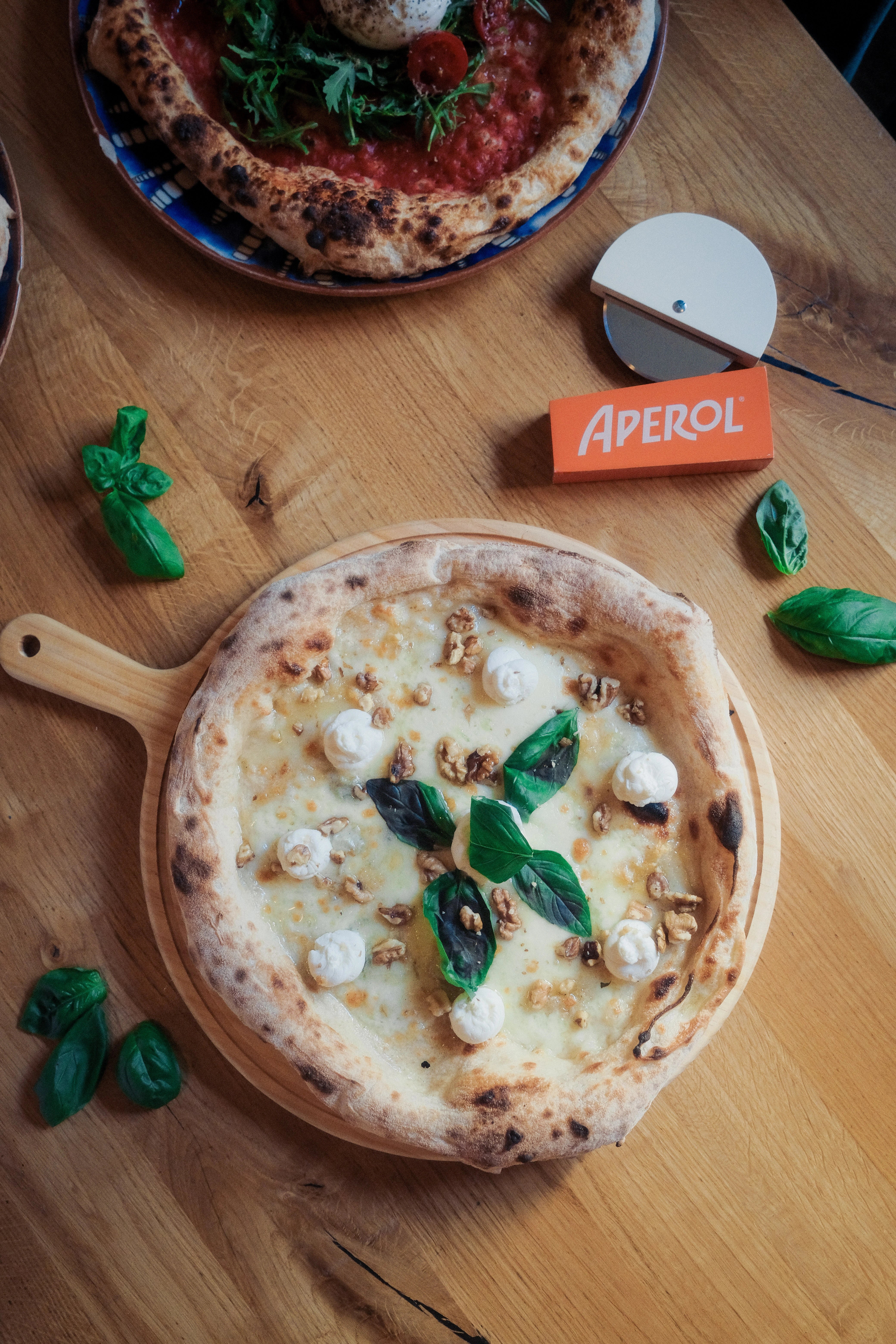 Small pizza with cheese and basil on a wooden board, with an Aperol sign in the background.