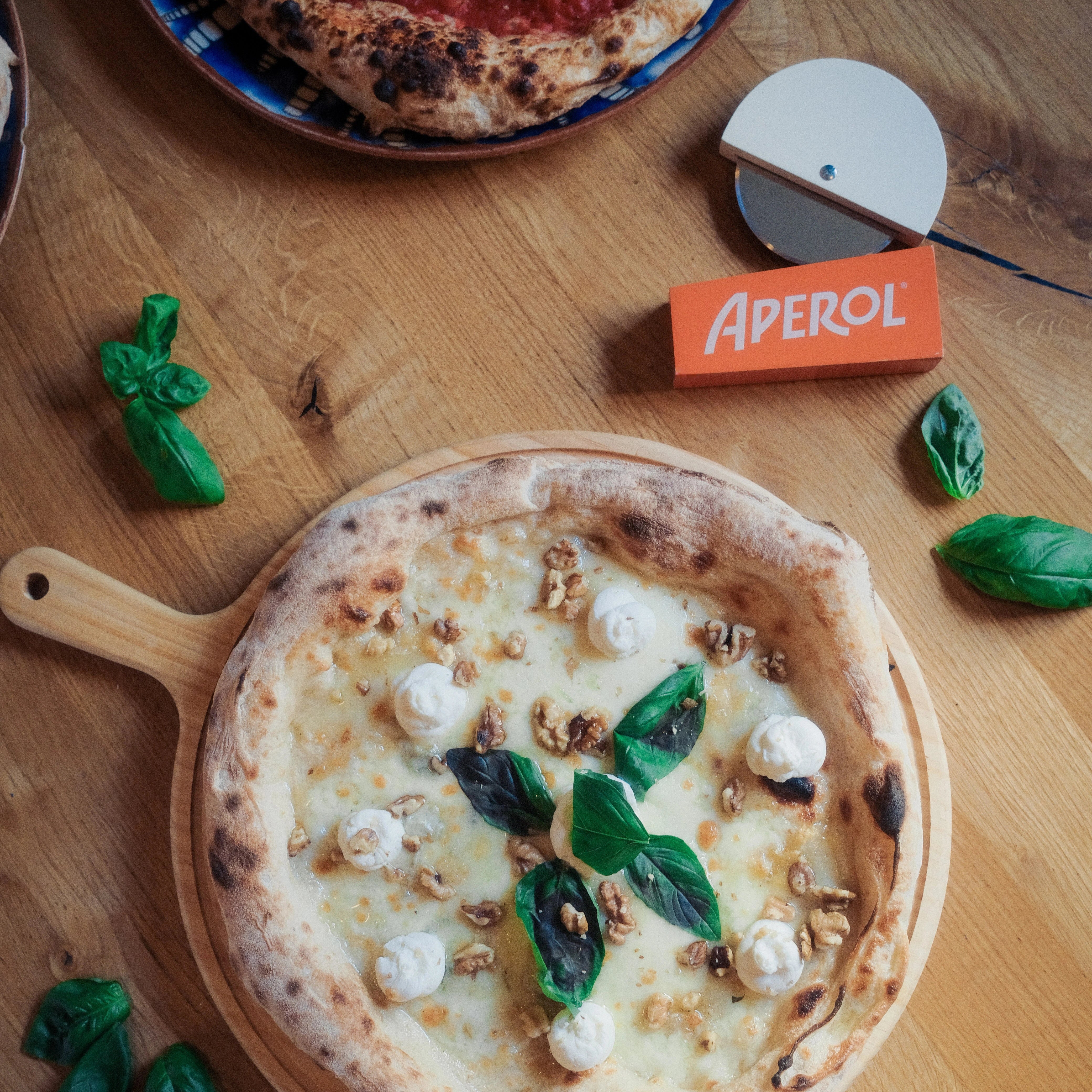 Two pizzas on a wooden table with Aperol box and pizza cutter