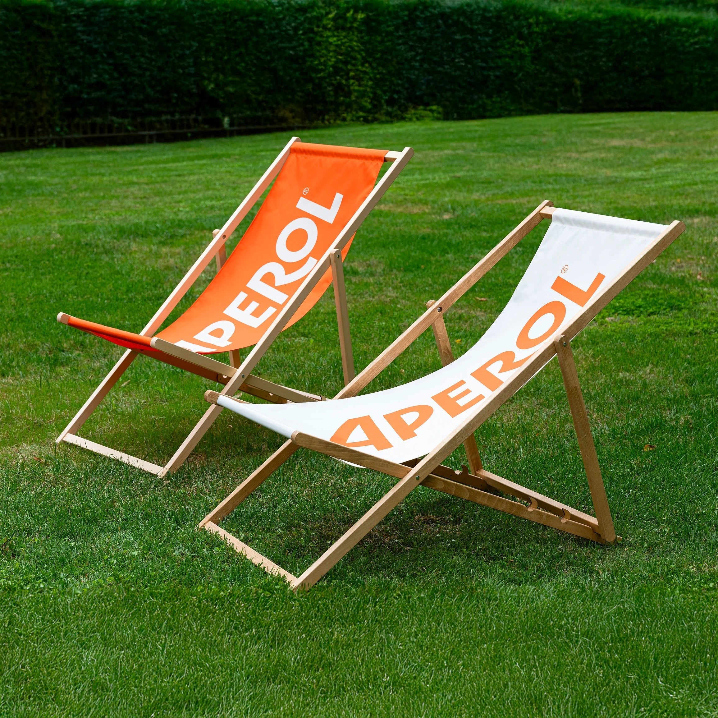 Two Aperol wooden deck chairs on a green lawn: one with a white fabric, the other orange, both featuring the bold Aperol logo, set in a leafy garden.