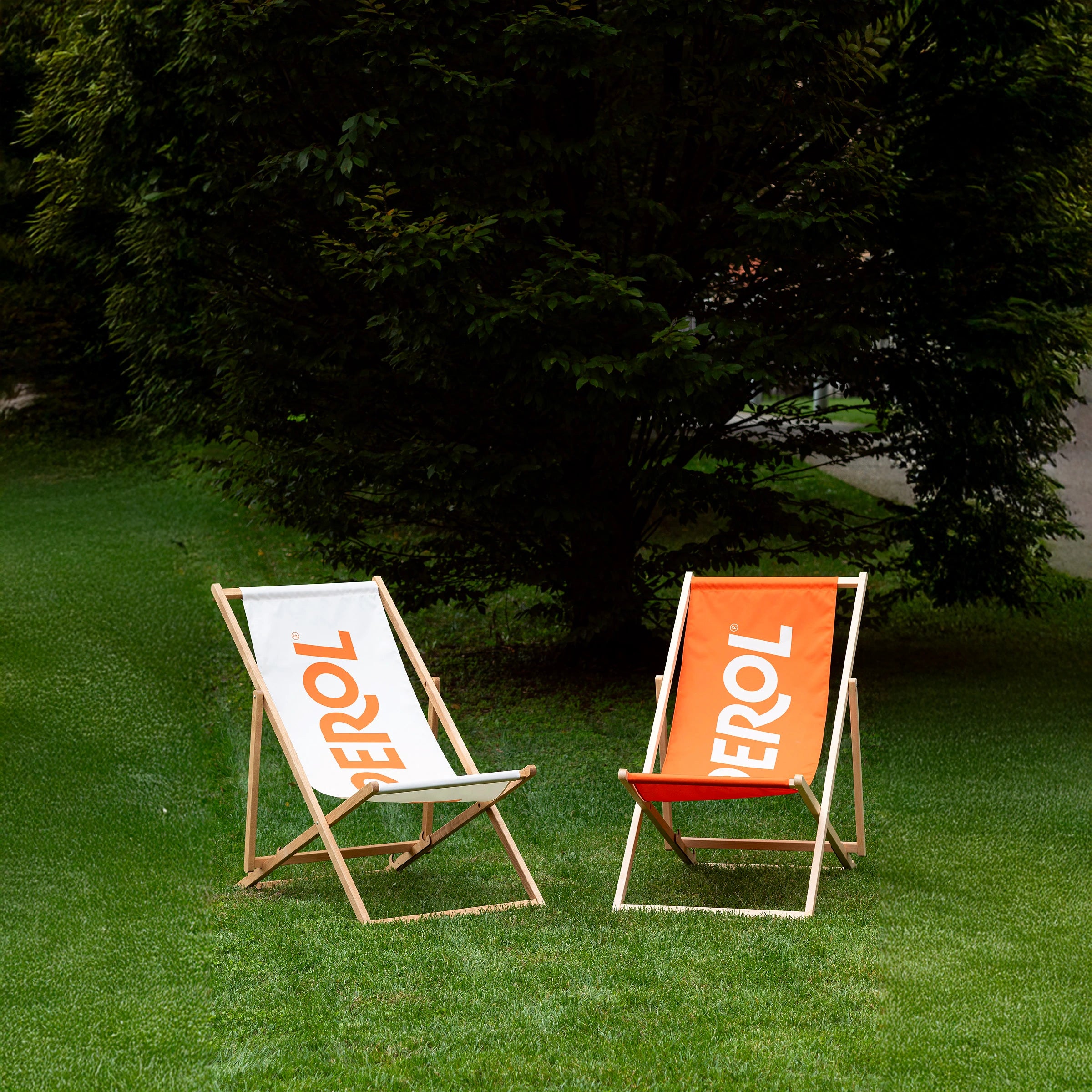 Two Aperol wooden deck chairs on a green lawn: one with a white fabric, the other orange, both featuring the bold Aperol logo, set in a leafy garden.