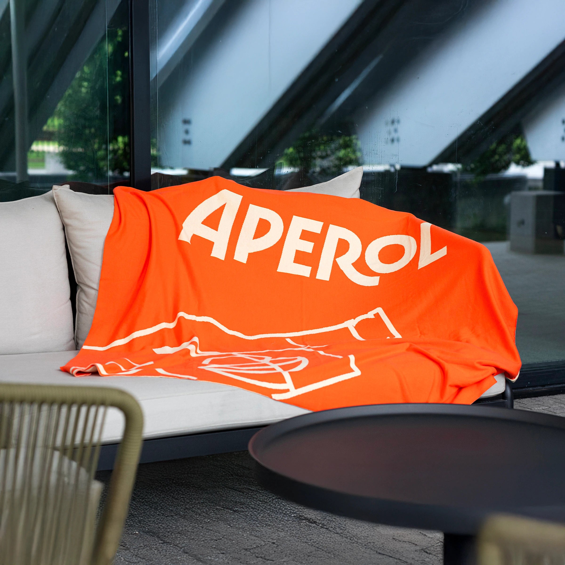 Bright orange Aperol winter blanket with white logo and cocktail glass graphic, draped over an outdoor sofa.