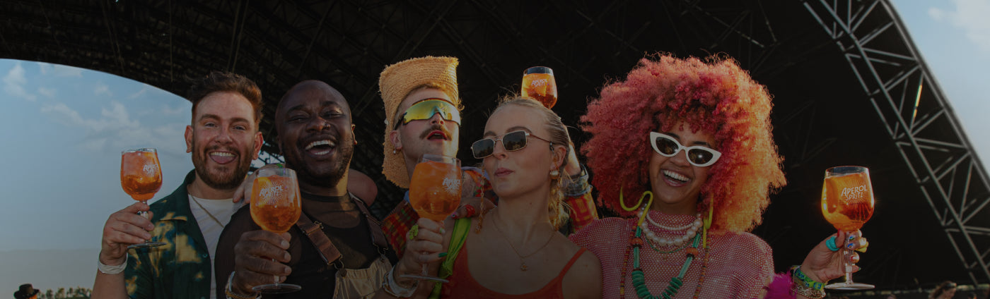 Group of five friends smiling and raising Aperol Spritz glasses in front of a large festival stage, enjoying a vibrant summer atmosphere.