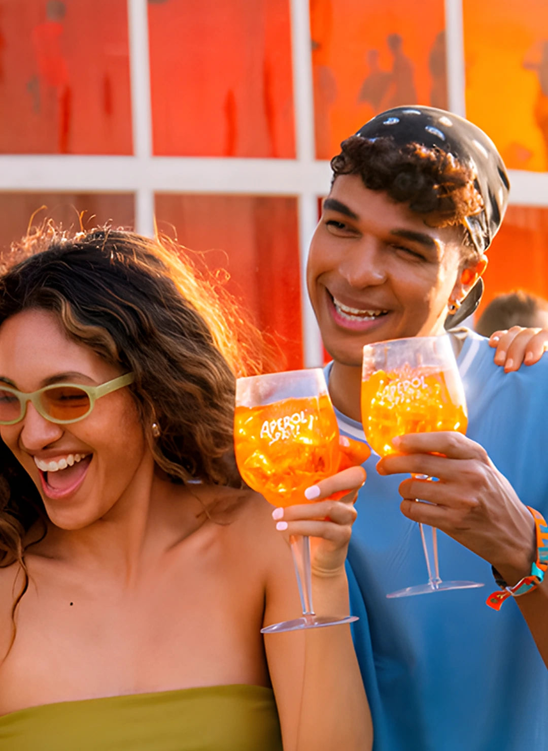 Two smiling friends clinking Aperol Spritz glasses at a sunny outdoor event, radiating a vibrant and summery atmosphere.