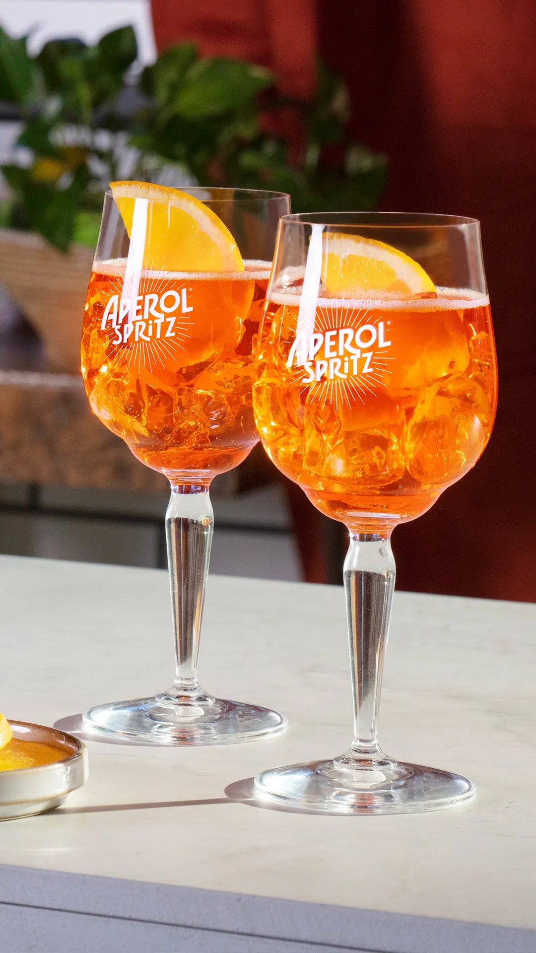 Two Aperol Spritz cocktails served in branded stem glasses with orange slices and ice cubes, placed on a bright countertop with a blurred background of greenery and warm-toned decor.