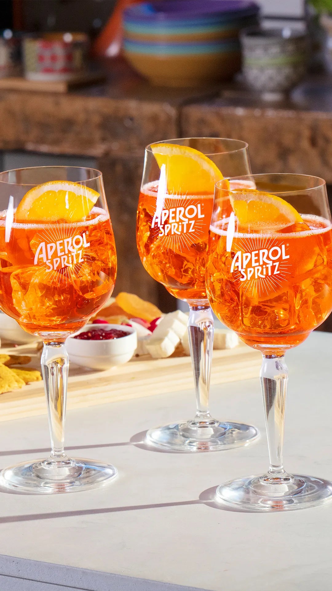 Three Aperol Spritz cocktails served in branded stem glasses, each garnished with a slice of orange and filled with ice, set on a white tabletop with a wooden board of snacks in the background.