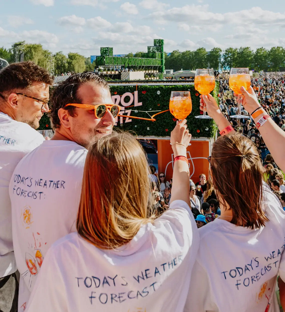 Group of friends at an Aperol Spritz festival, wearing matching shirts that read 'Today's Weather Forecast', toasting with Aperol cocktails in front of a large crowd and branded bar setup under a sunny sky.