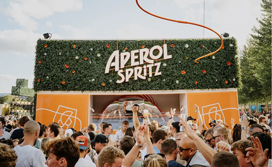 Large crowd enjoying a vibrant outdoor event in front of an Aperol Spritz booth decorated with greenery and orange accents at a summer festival.