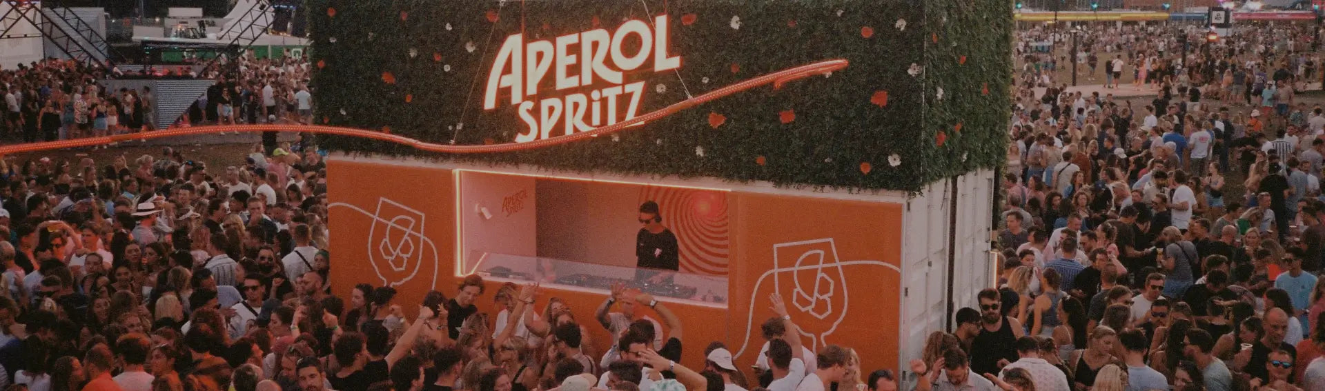 Massive crowd enjoying a sunny outdoor festival with Aperol Spritz-branded bars decorated with greenery and orange accents, set against a backdrop of trees and tents.
