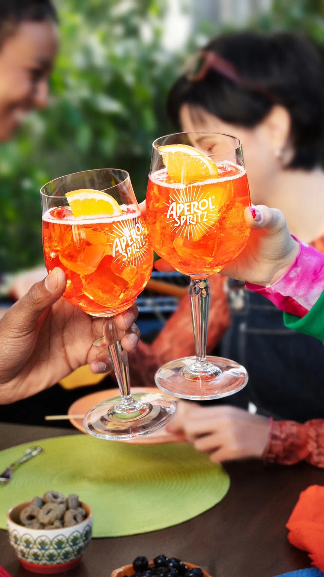 Two people toasting with Aperol Spritz cocktails in branded glasses, garnished with orange slices, while seated at a colorful outdoor table with snacks and drinks, surrounded by friends enjoying the moment.
