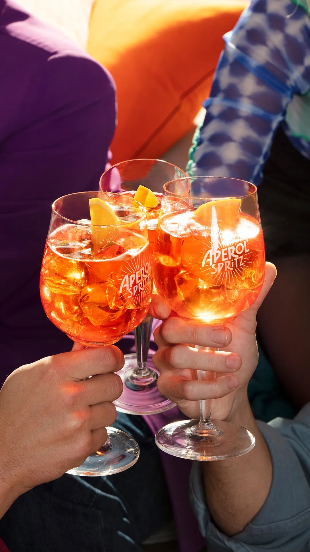 Close-up of three hands toasting with Aperol Spritz cocktails in branded glasses, garnished with orange slices, against a vibrant backdrop featuring colorful clothing and cushions, capturing a festive and cheerful atmosphere.