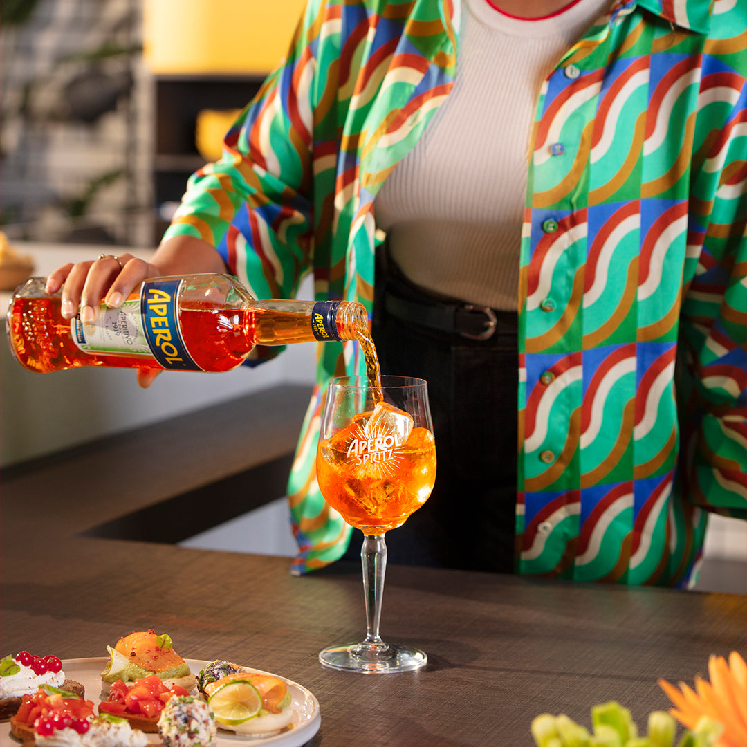 Person in a colorful patterned shirt pouring Aperol into a branded Aperol Spritz glass filled with ice, with a plate of assorted appetizers and garnishes on the table in the foreground, capturing a vibrant moment of drink preparation.