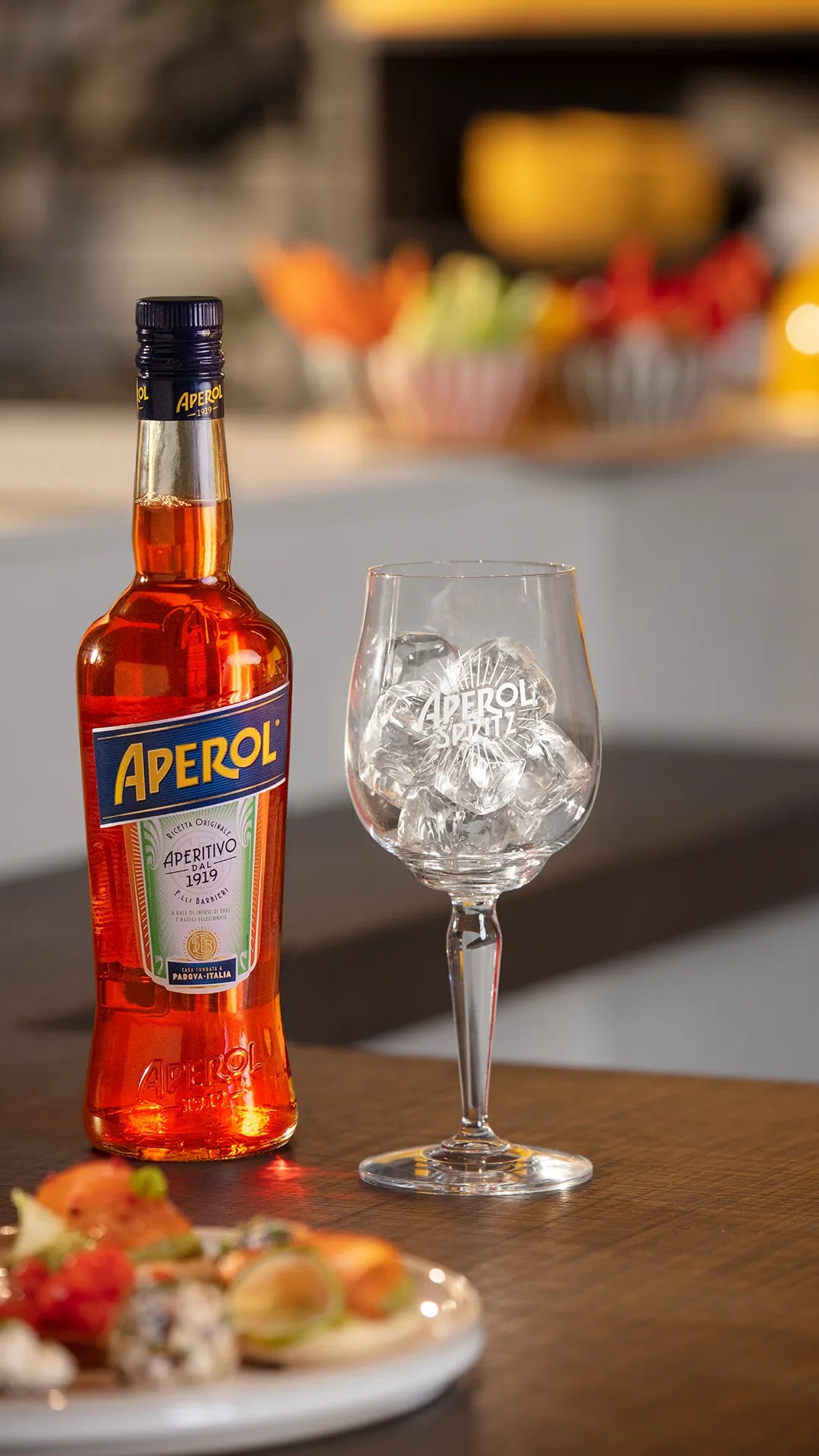 A bottle of Aperol stands beside a branded wine glass filled with ice cubes on a wooden countertop, ready for a refreshing cocktail. In the foreground, a plate of colorful appetizers adds to the inviting aperitivo atmosphere.