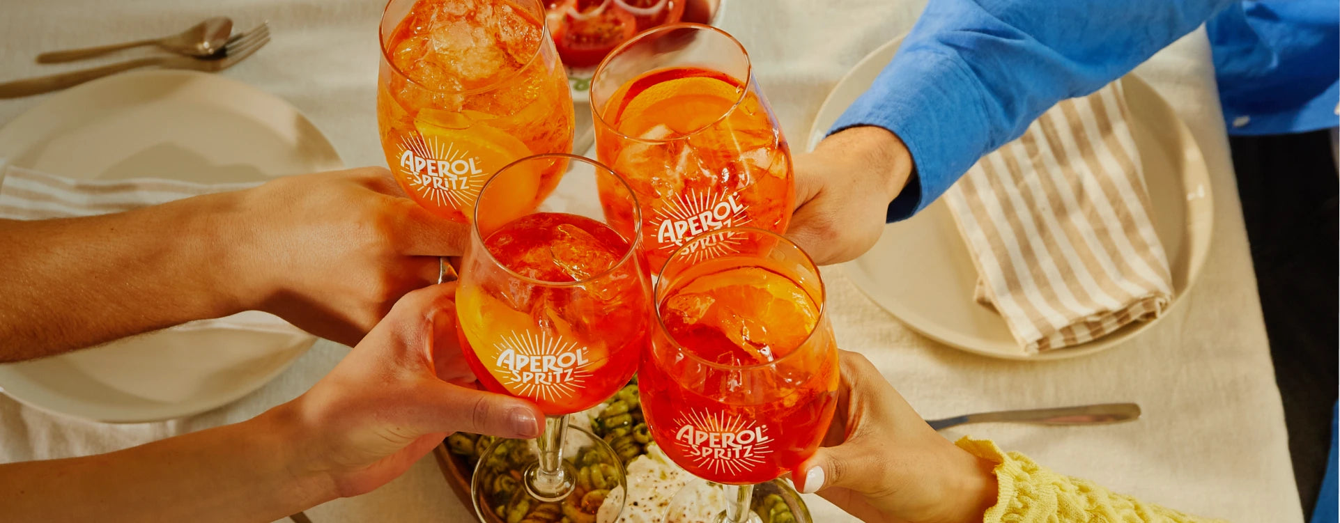 Four people clinking Aperol Spritz glasses over a festive table set with plates, cutlery, and appetizers.
