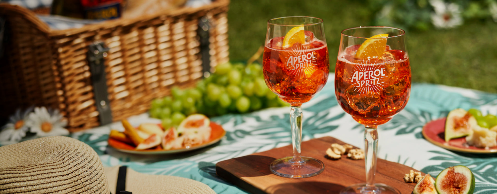 Two Aperol Spritz glasses on a wooden board, placed on a tropical tablecloth. In the background: a picnic basket, green grapes, sliced figs, cheese, a straw hat, and white flowers on the grass.