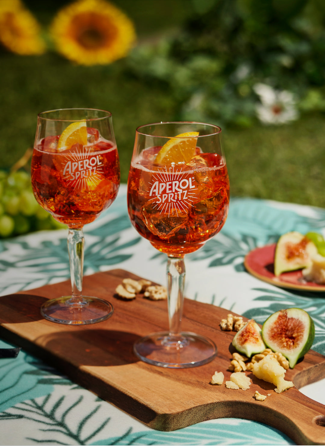 Two glasses of Aperol Spritz on a wooden board, served with fresh figs, walnuts, and crumbled cheese, set on a tropical-themed tablecloth outdoors with sunflowers in the background.