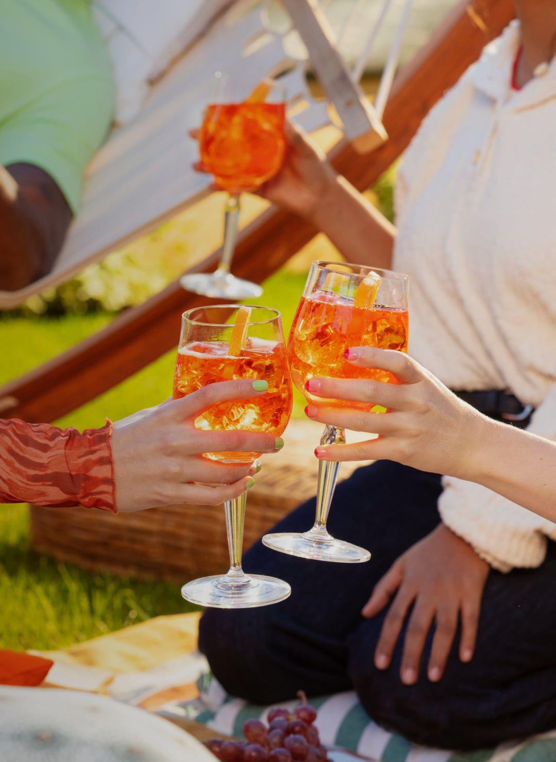 Three glasses of Aperol Spritz being clinked together during a picnic on the grass, with people seated nearby, a hammock in the background, and grapes in the foreground.