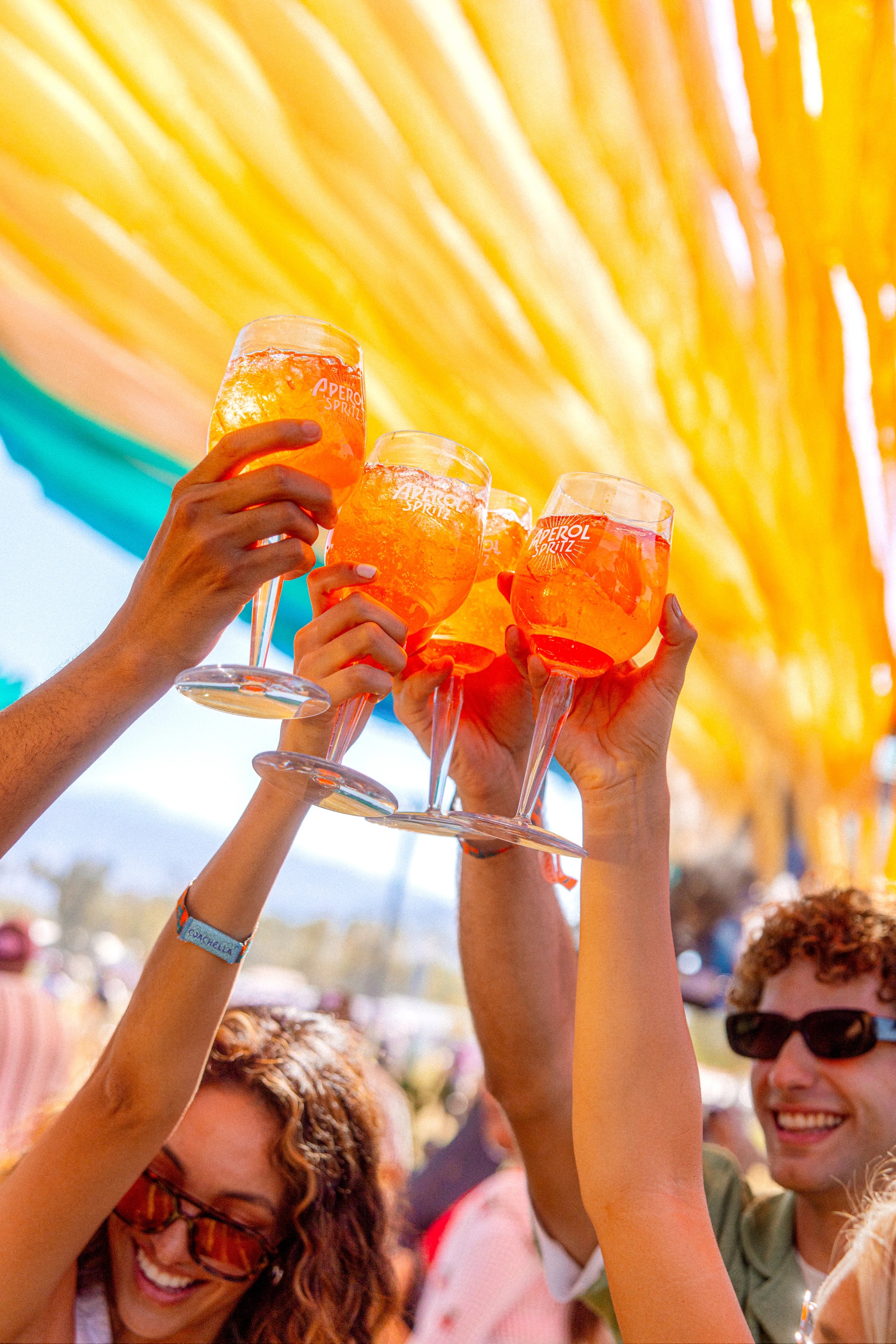 Friends cheerfully toasting with raised Aperol Spritz glasses beneath a canopy of yellow streamers, capturing a vibrant and sunny outdoor celebration.