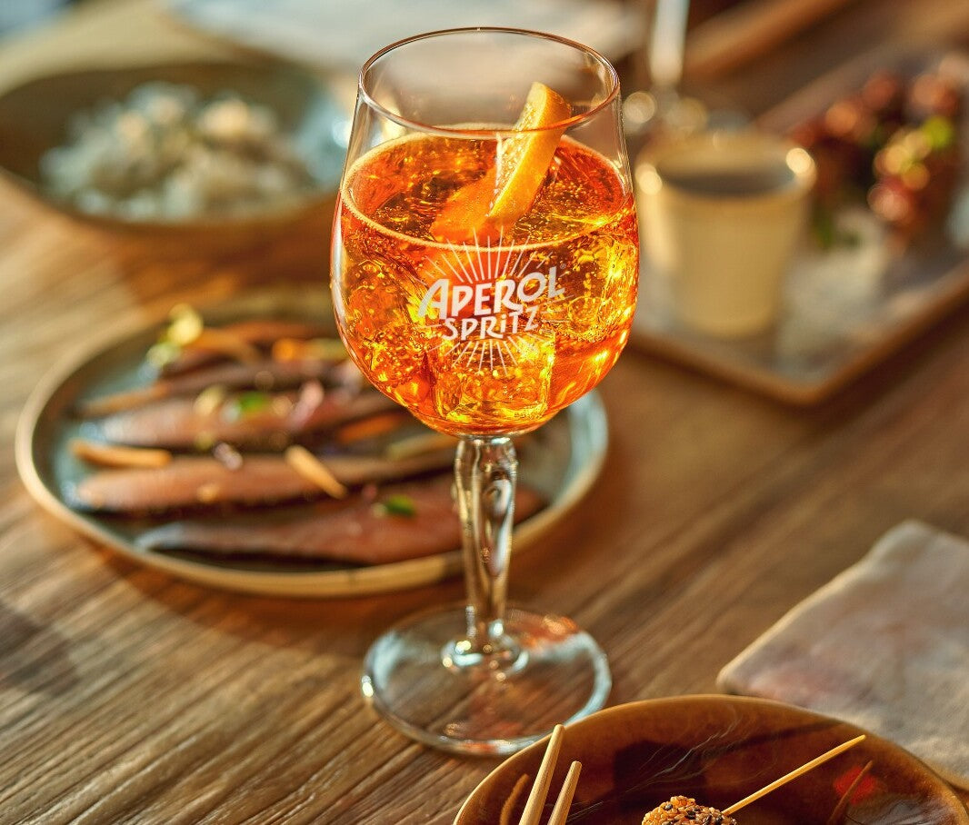 A close-up of a sparkling Aperol Spritz served in a branded glass with an orange slice garnish, placed on a rustic wooden table surrounded by assorted appetizers and finger foods.