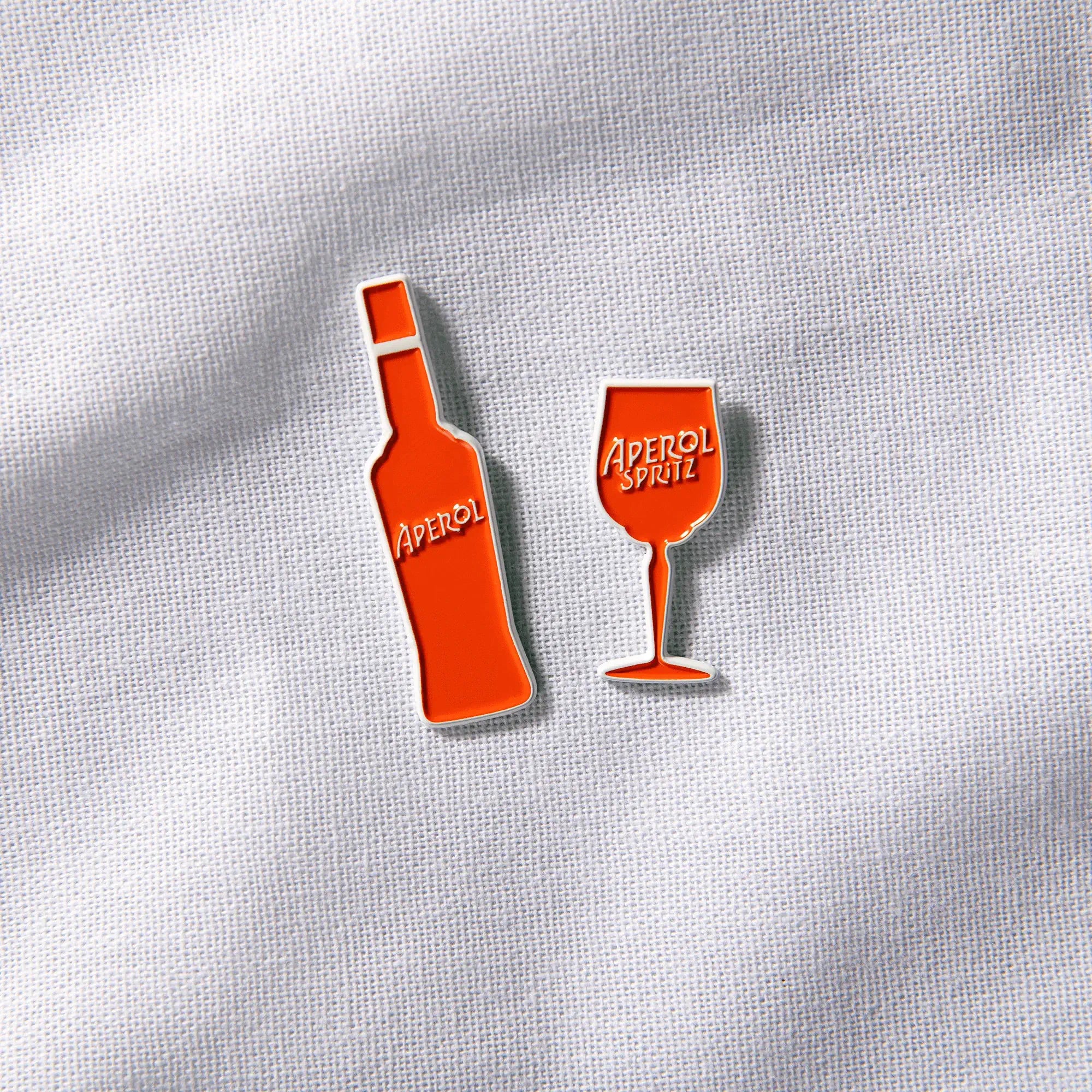 Two enamel pins in the shape of an Aperol bottle and a wine glass with the Aperol Spritz logo, displayed on a textured white fabric background.