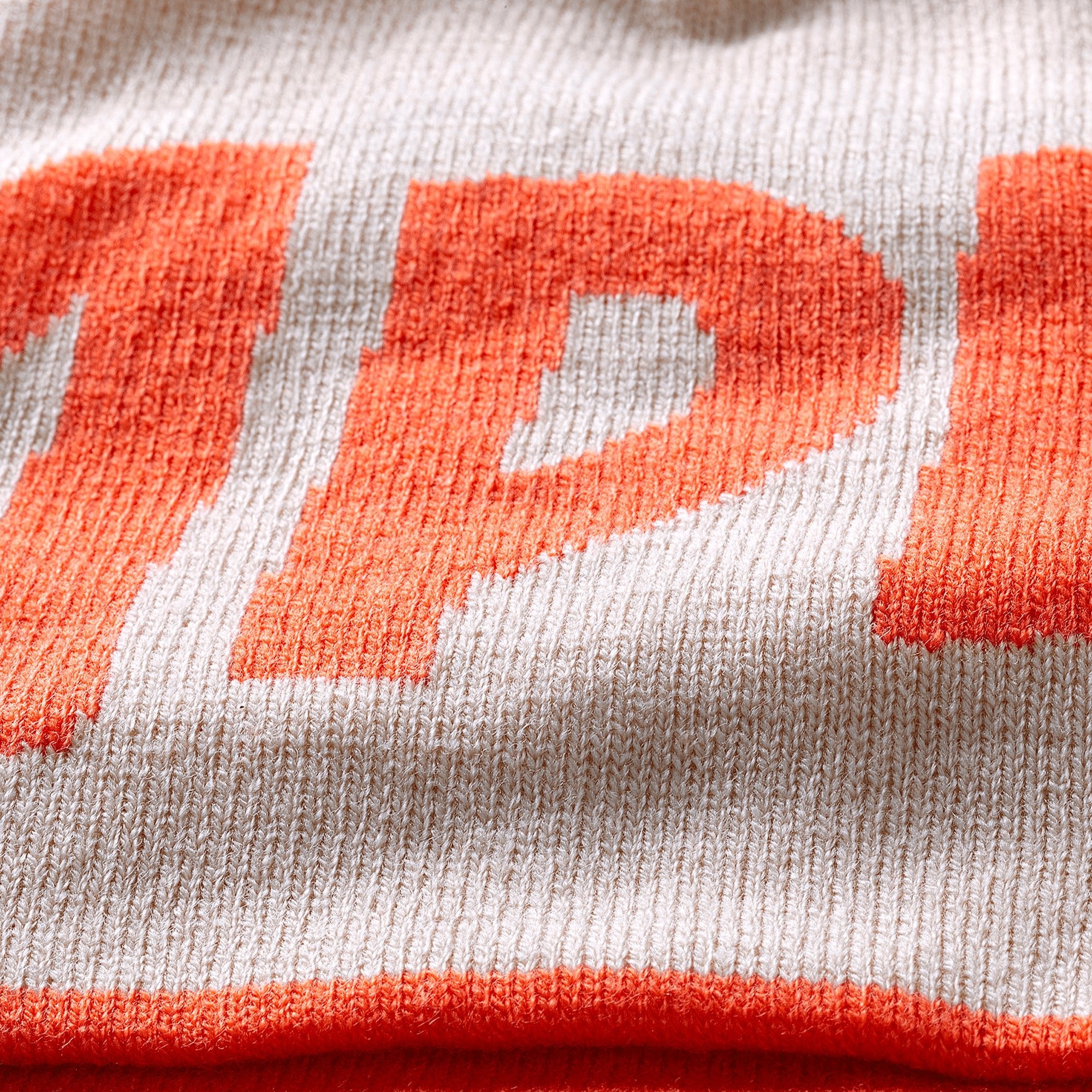 Close-up of a beige and orange knitted winter hat, showing the orange letter “P” as part of the word “APEROL”.