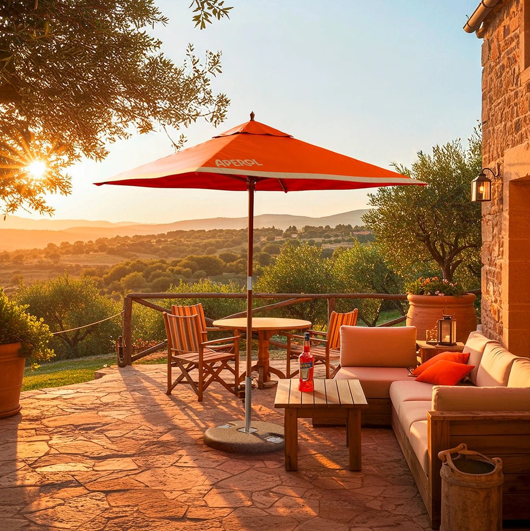 Scenic outdoor terrace at sunset featuring an Aperol-branded orange parasol, cozy wooden furniture, a bottle of Aperol on the table, and a panoramic view of rolling hills and olive trees under a golden sky.