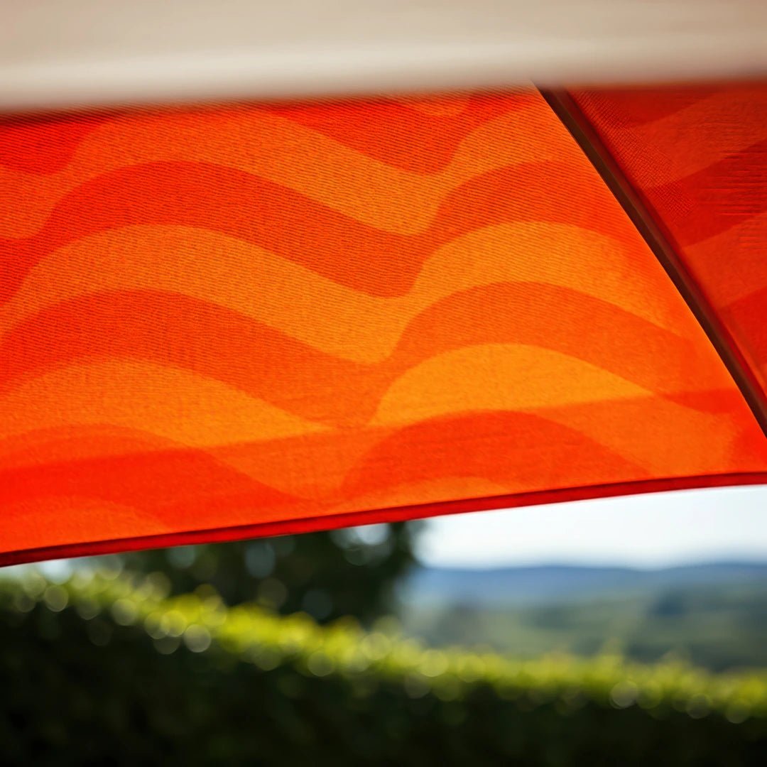 Close-up upward view of an orange wavy-patterned parasol with sunlight filtering through, set against a blurred background of greenery and rolling hills, evoking a warm and summery atmosphere.
