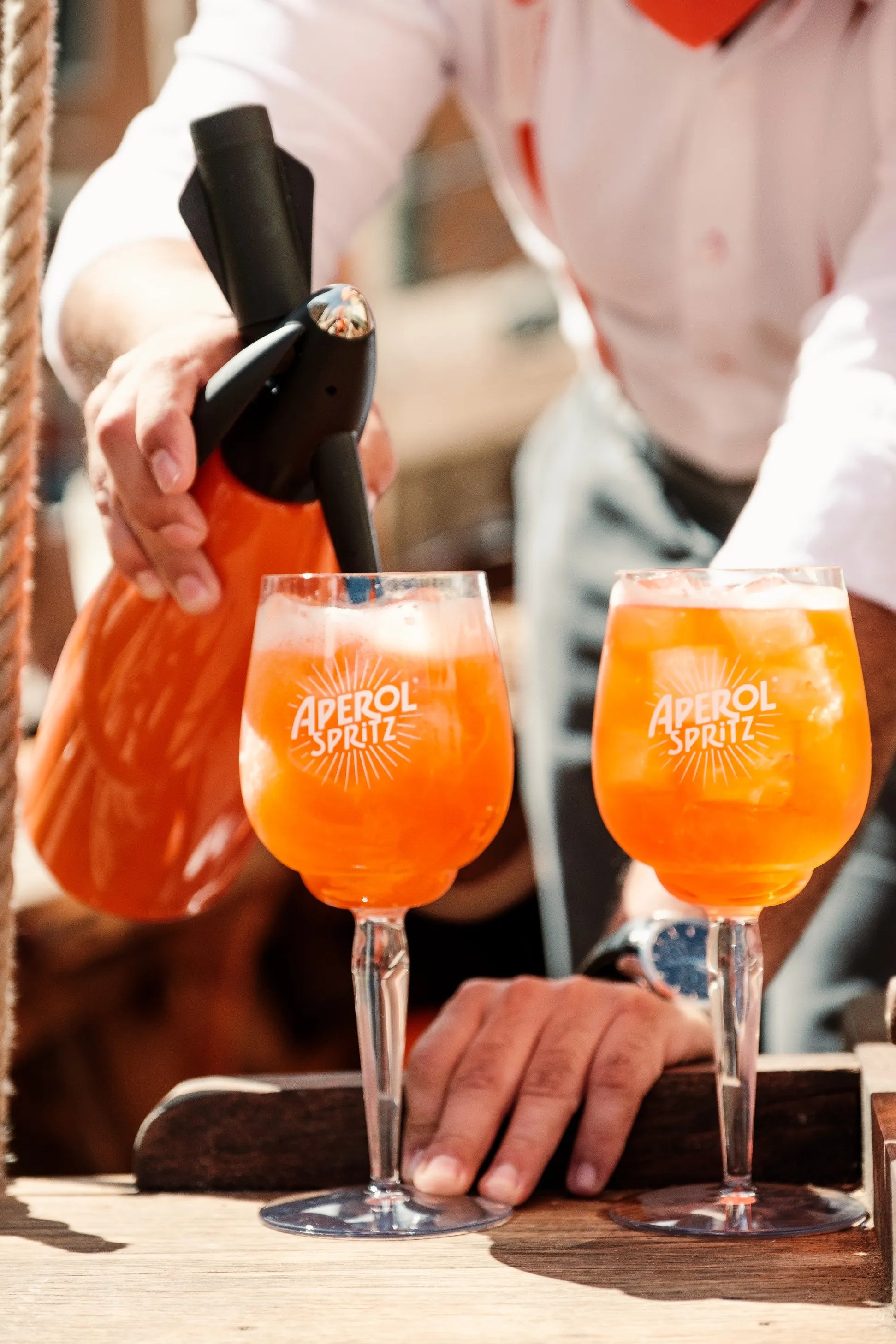 A bartender pours soda water into an Aperol Spritz using an orange soda siphon, with two branded Aperol glasses filled with ice and the signature orange cocktail resting on a wooden counter.