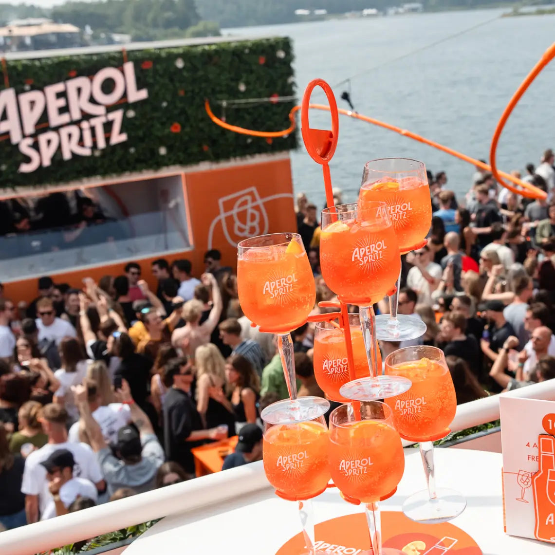 A festive outdoor Aperol Spritz event by the water, with a lively crowd in the background and a stand showcasing several vibrant Aperol Spritz cocktails in the foreground.