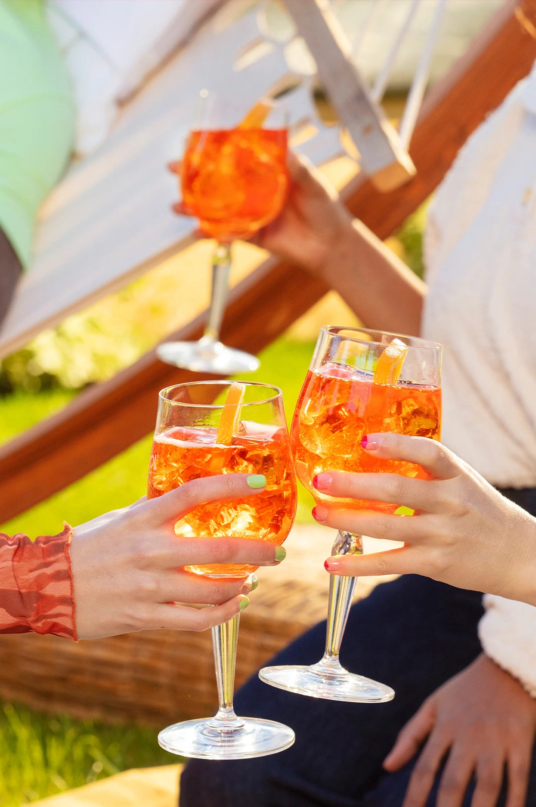 Three people clinking Aperol Spritz glasses filled with ice and orange slices, enjoying a sunny outdoor moment together.