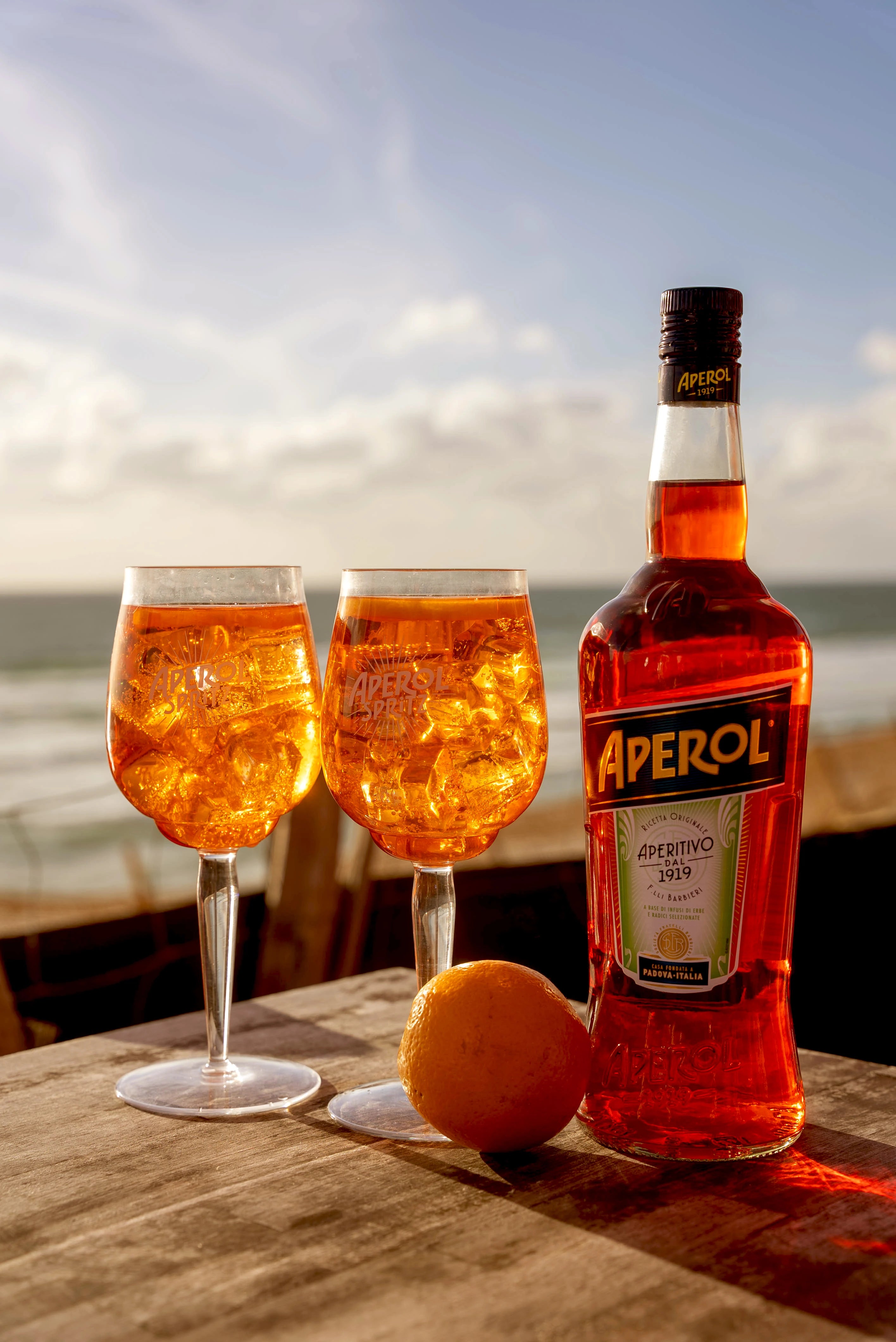 Bottle of Aperol with orange and two Aperol Spritz cocktails on wooden table by the sea at sunset