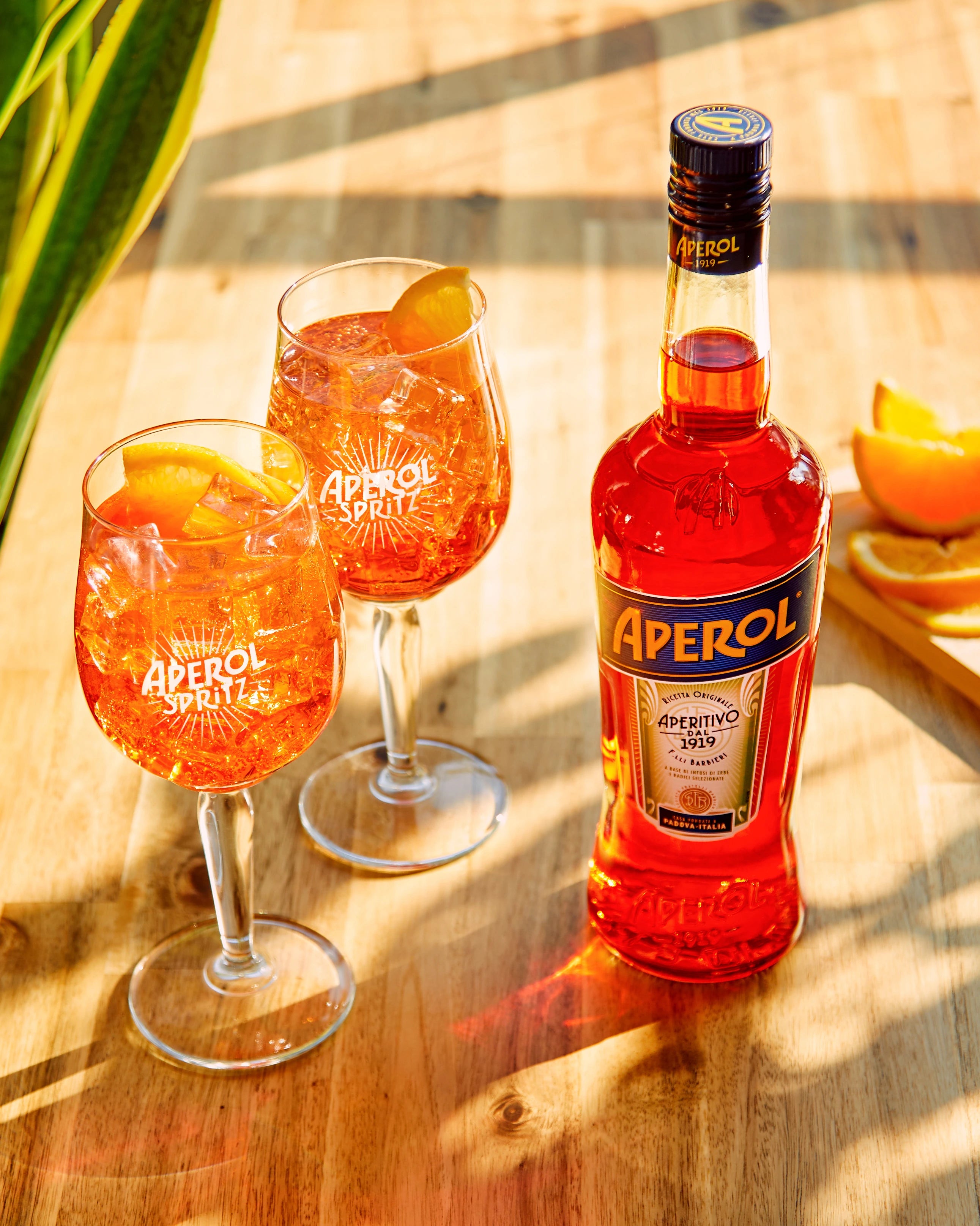 Two Aperol Spritz glasses with ice and orange slices on a sunlit wooden table, next to an Aperol bottle and a few orange wedges.