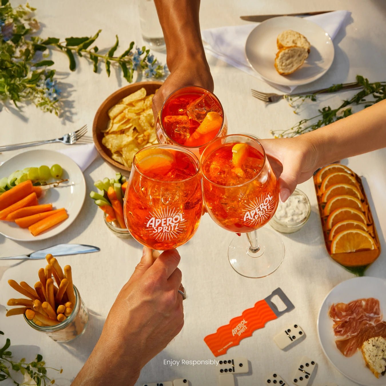 Three Aperol Spritz glasses clinking over a summer table with snacks, vegetables, bread and dominoes
