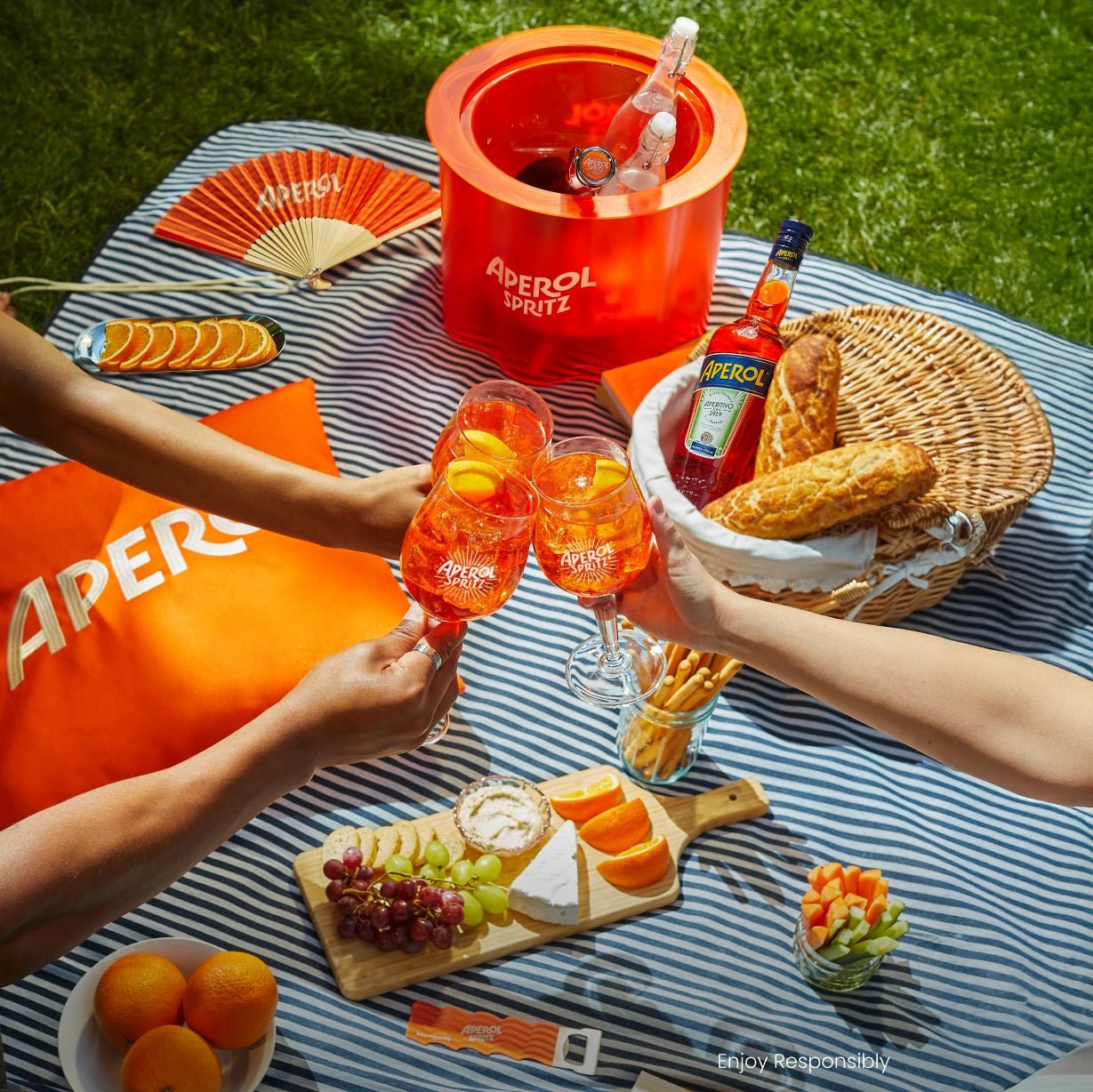 Hands toasting Aperol Spritz at a summer picnic with striped blanket, snacks, fan, and branded cooler