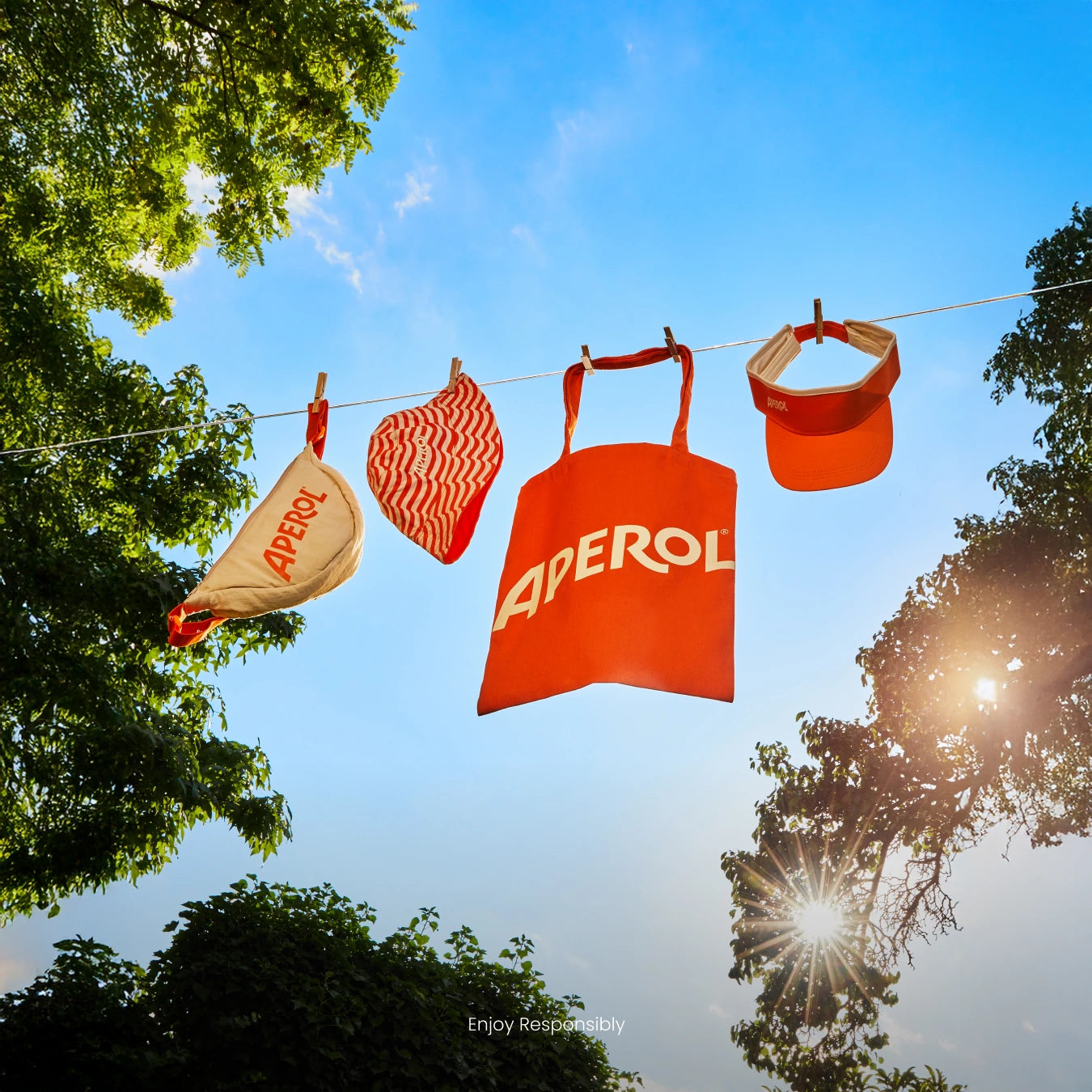 Aperol tote bag, pouch, and visor hanging on a clothesline in the sun, surrounded by trees and blue sky