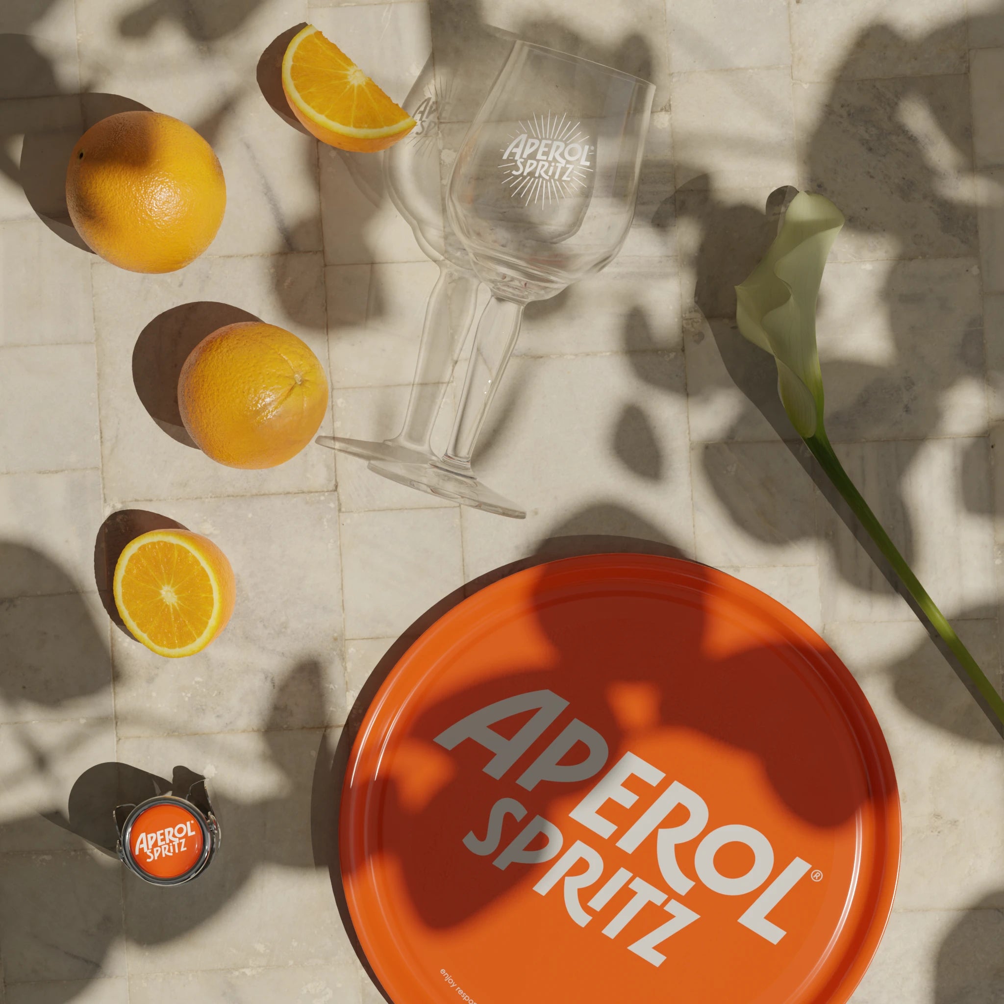 Aperol Spritz glasses, oranges, branded tray, and bottle cap arranged on sunlit stone surface with floral shadow