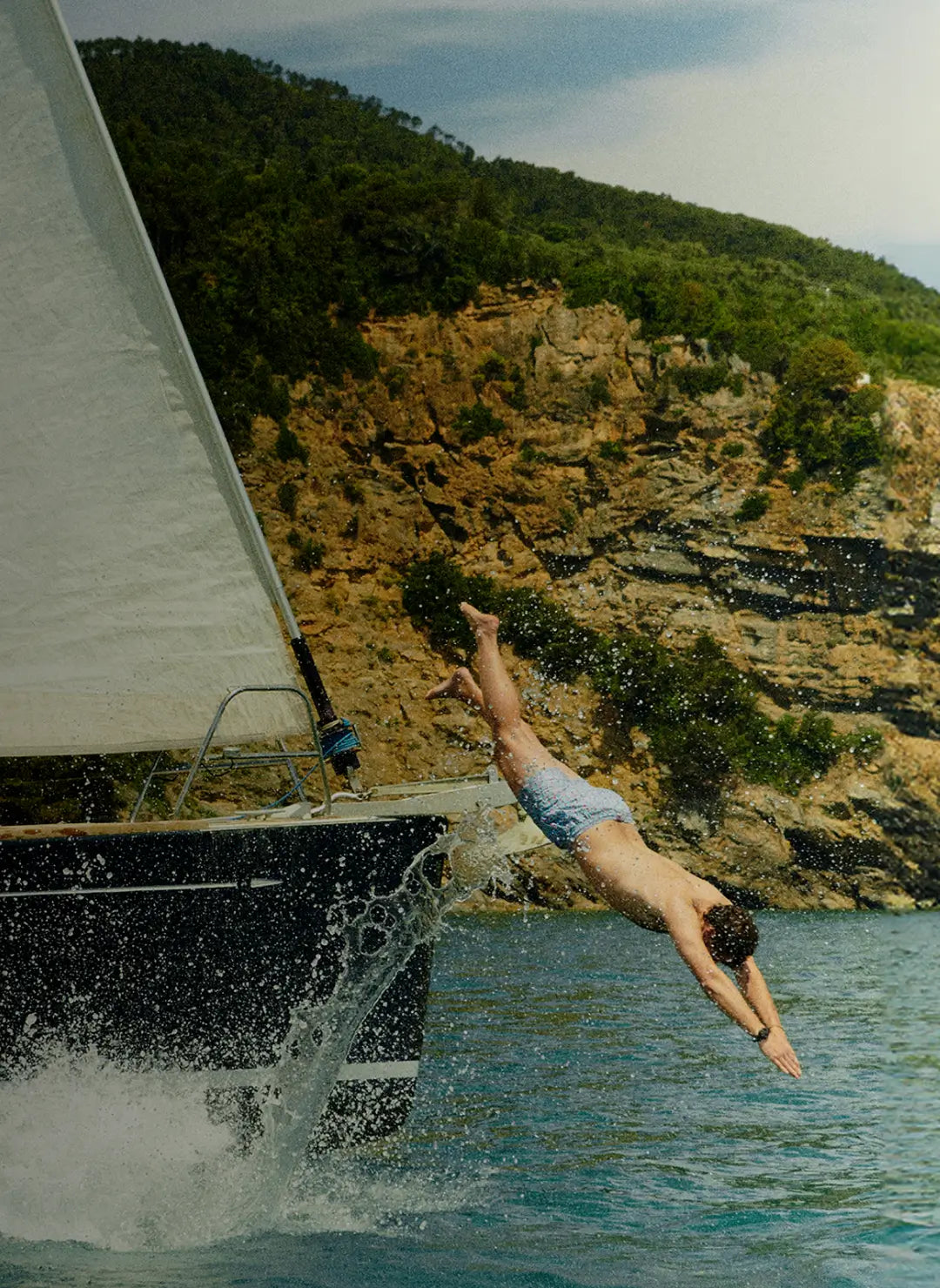 Man diving from a sailboat into turquoise sea, with a rocky, green cliffside in the background.