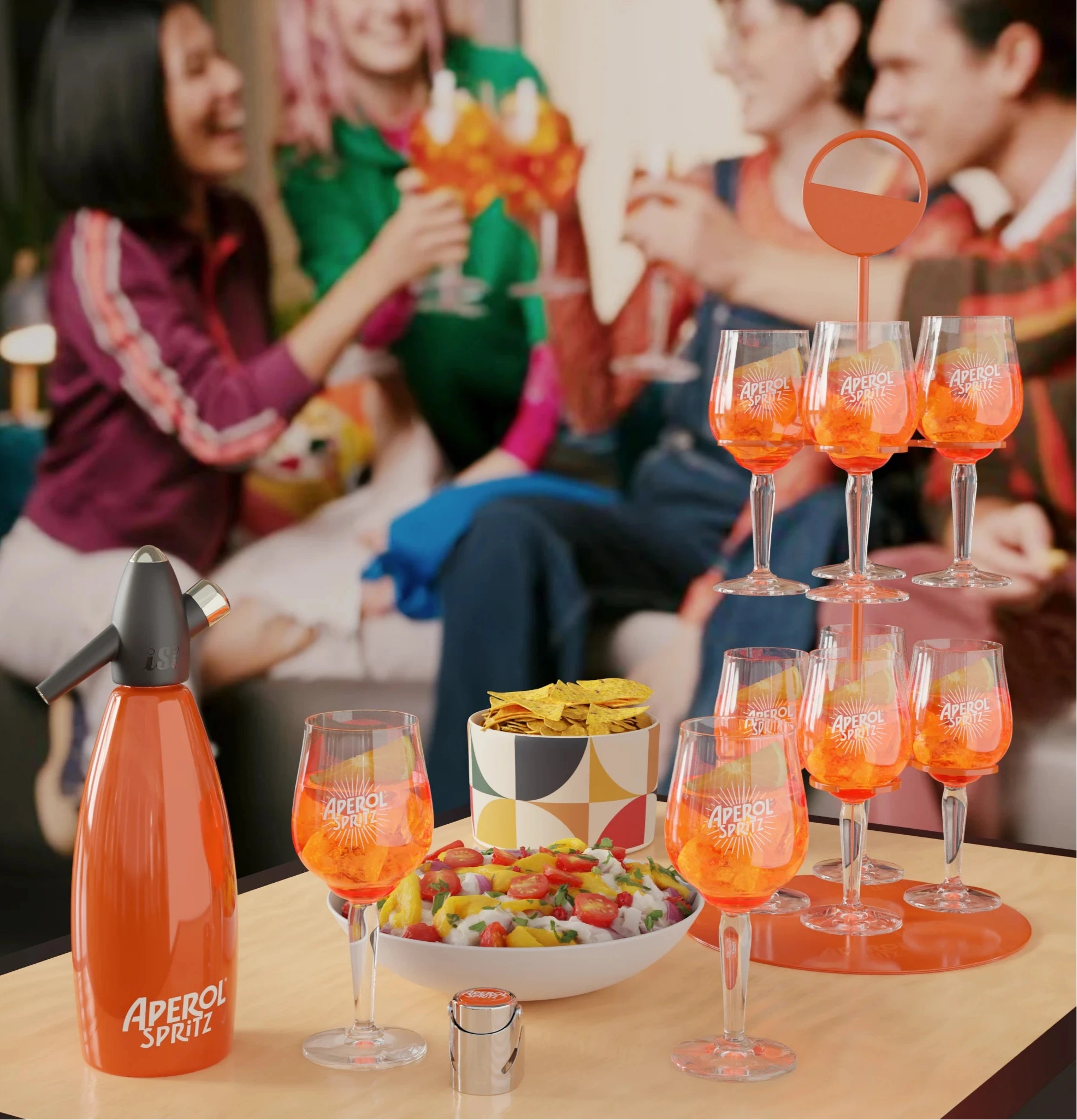 A lively indoor setting with a group of friends enjoying Aperol Spritz together. In the foreground, a two-tiered orange Aperol Spritz group tray stand displays several filled glasses on a wooden table. The table also holds a bowl of tortilla chips, a fruit salad, a branded Aperol Spritz siphon, and a few extra glasses. The background shows friends smiling and raising their glasses, creating a warm and festive atmosphere.