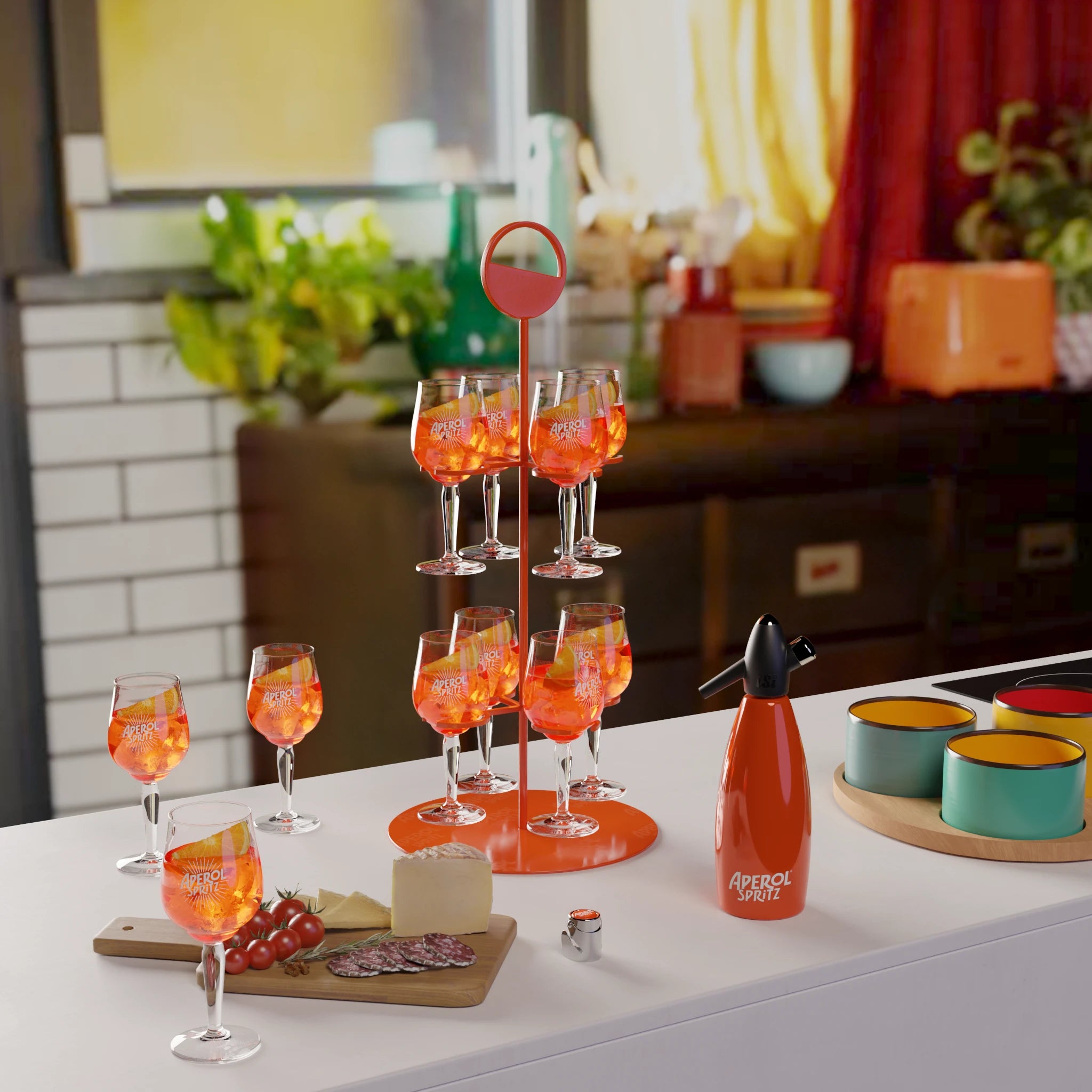 A vibrant kitchen scene featuring a two-tiered orange Aperol Spritz group tray stand, holding multiple filled glasses. Surrounding the tray are additional Aperol Spritz glasses, a cutting board with cheese, salami, and cherry tomatoes, a branded Aperol Spritz siphon, and colorful bowls, all set on a modern white kitchen island.