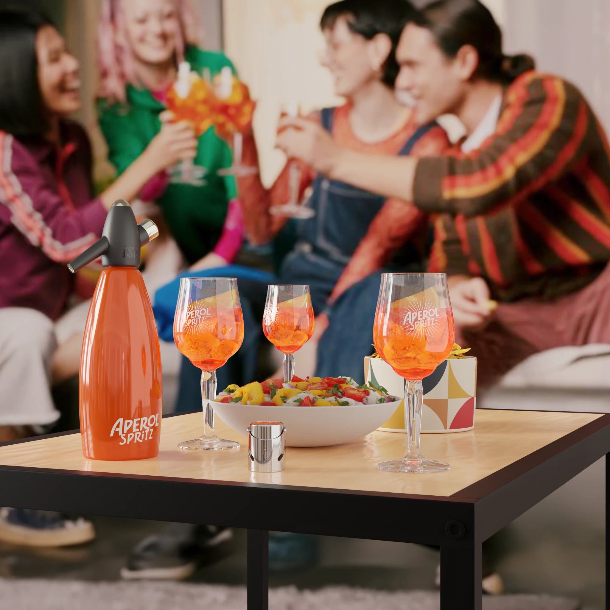 A group of friends cheerfully toasting in a living room. In the foreground, a coffee table displays three Aperol Spritz glasses, an orange siphon labeled "Aperol Spritz", a colorful salad bowl, and a stylish snack box.