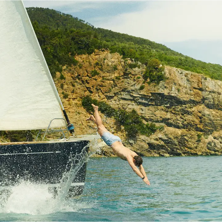 Man diving from a sailboat into turquoise sea, with a rocky, green cliffside in the background.