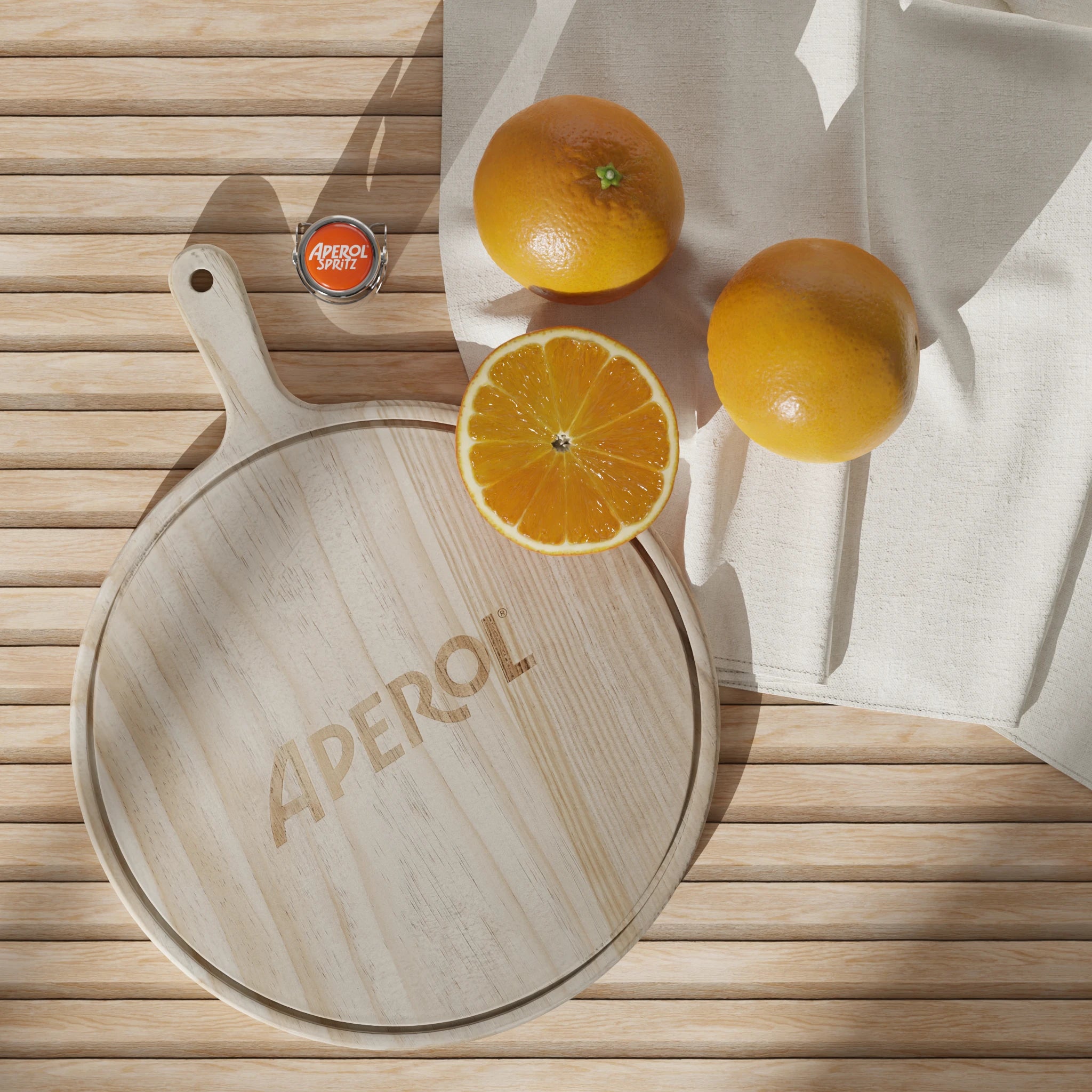 Top-down view of a round wooden Aperol-branded pizza tray on a slatted wooden surface, accompanied by two whole oranges, one halved orange, a white napkin, and an Aperol Spritz bottle cap.