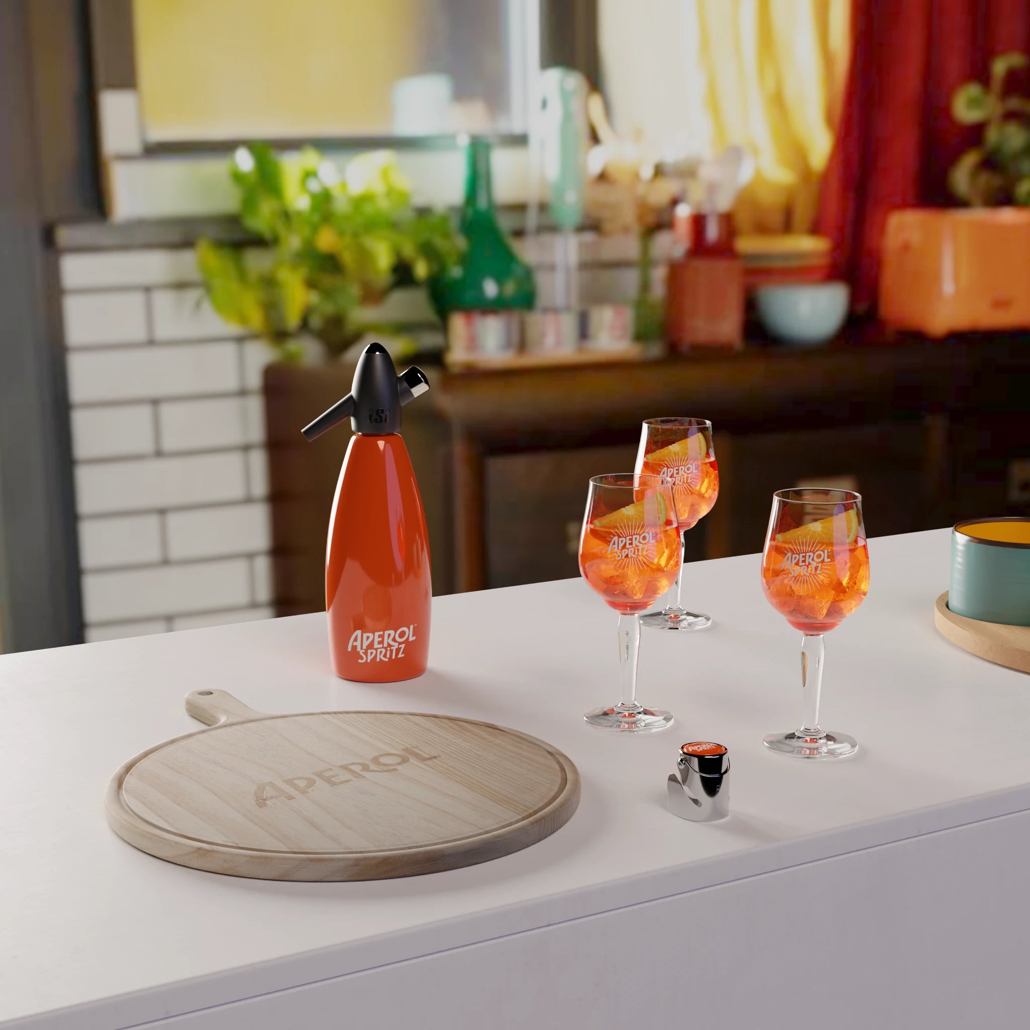 A kitchen counter featuring an Aperol-branded wooden pizza tray, a bright orange Aperol soda siphon, two Aperol Spritz glasses, and a bottle stopper. In the background, a cozy, colorful kitchen is visible with plants, utensils, and jars on shelves.