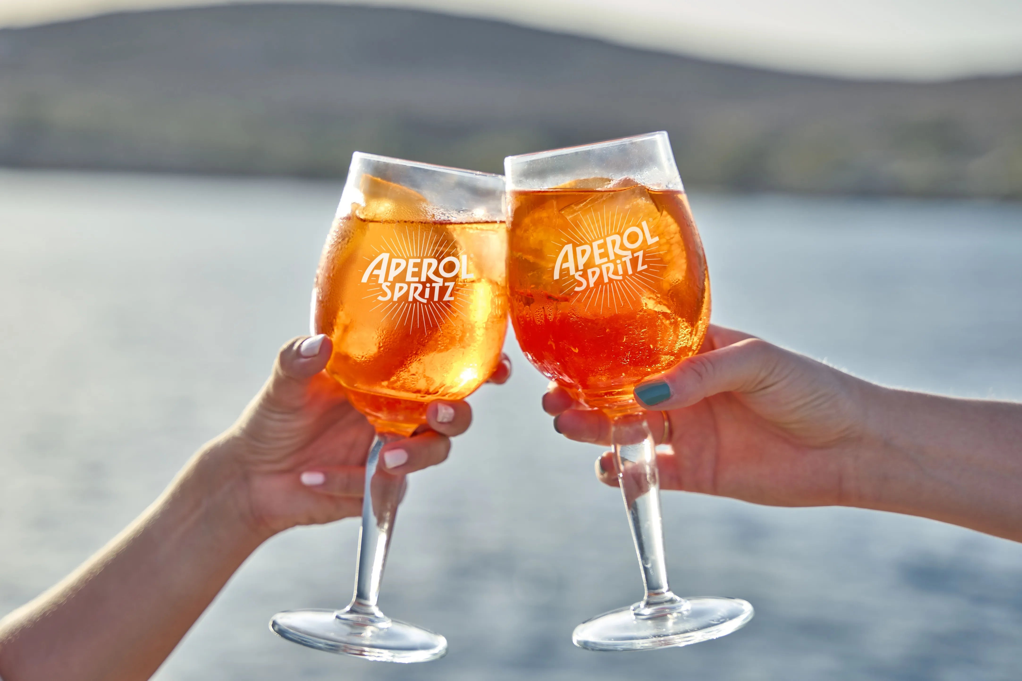 Two hands clinking Aperol Spritz glasses filled with ice and orange slices in front of a scenic waterfront view at sunset, celebrating the moment with a refreshing toast.