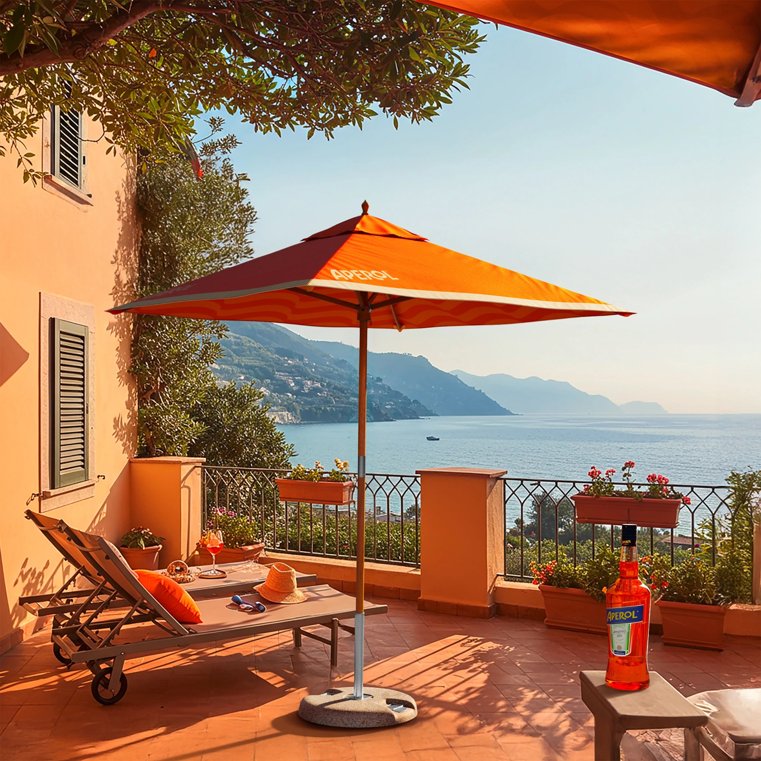 Sunny coastal terrace featuring an Aperol-branded orange parasol, lounge chairs with orange pillows, a bottle of Aperol on a small table, and a stunning sea view with distant mountains and a boat on calm water.