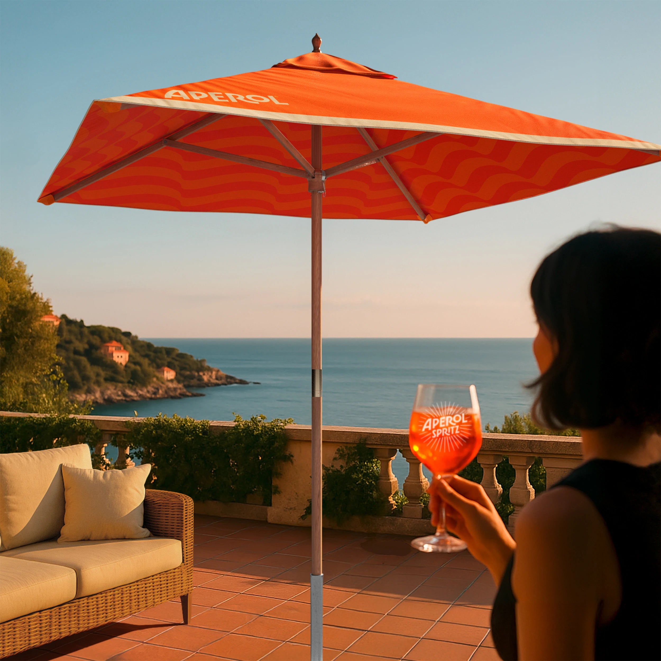 Woman holding an Aperol Spritz under a vibrant orange Aperol-branded parasol on a sunny seaside terrace, with a scenic view of the coastline and blue ocean in the background.